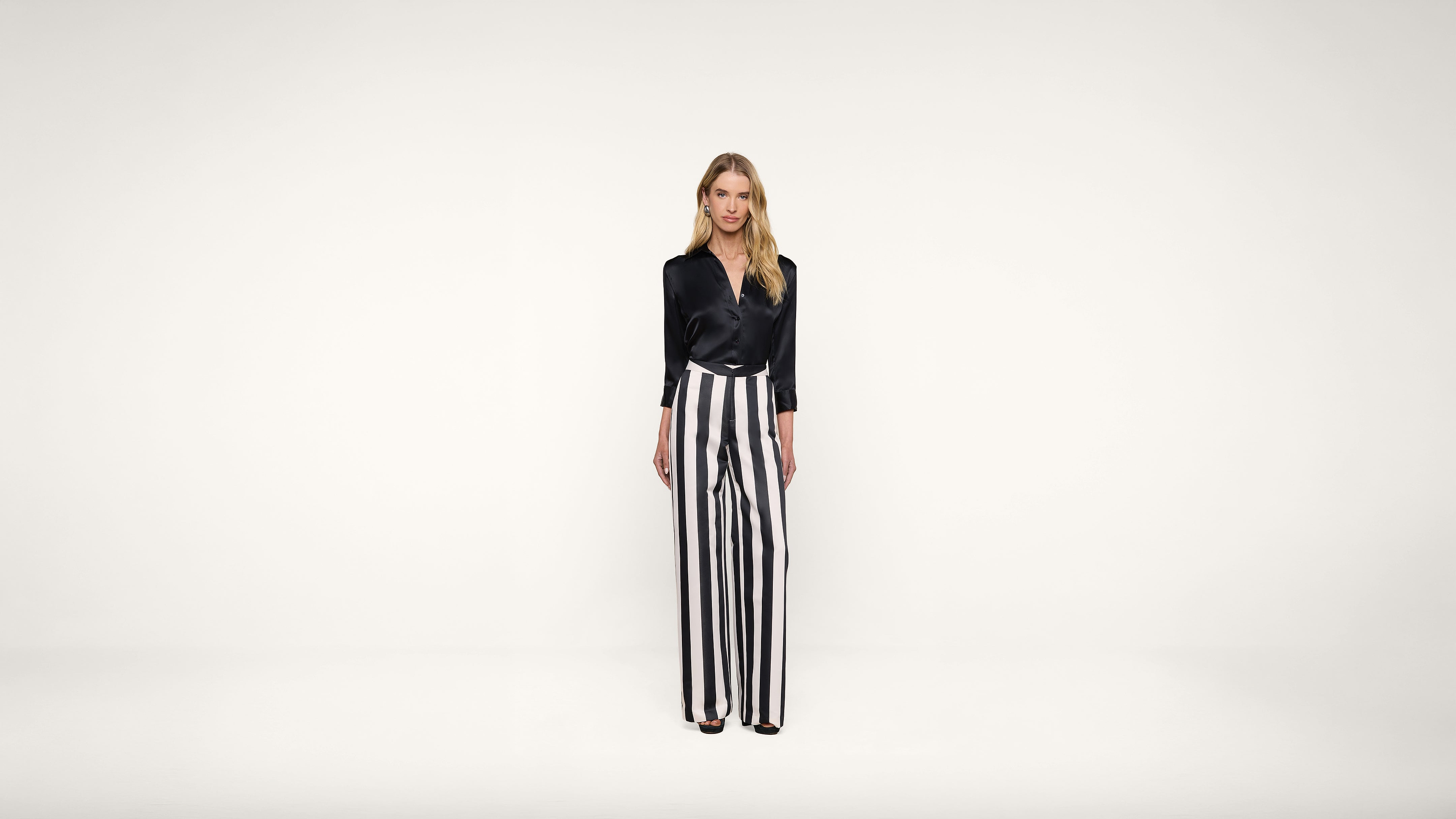 Woman stands against a plain white background wearing a black blouse and high-waisted, wide-leg black and white vertical striped pants. She faces forward with a neutral expression.