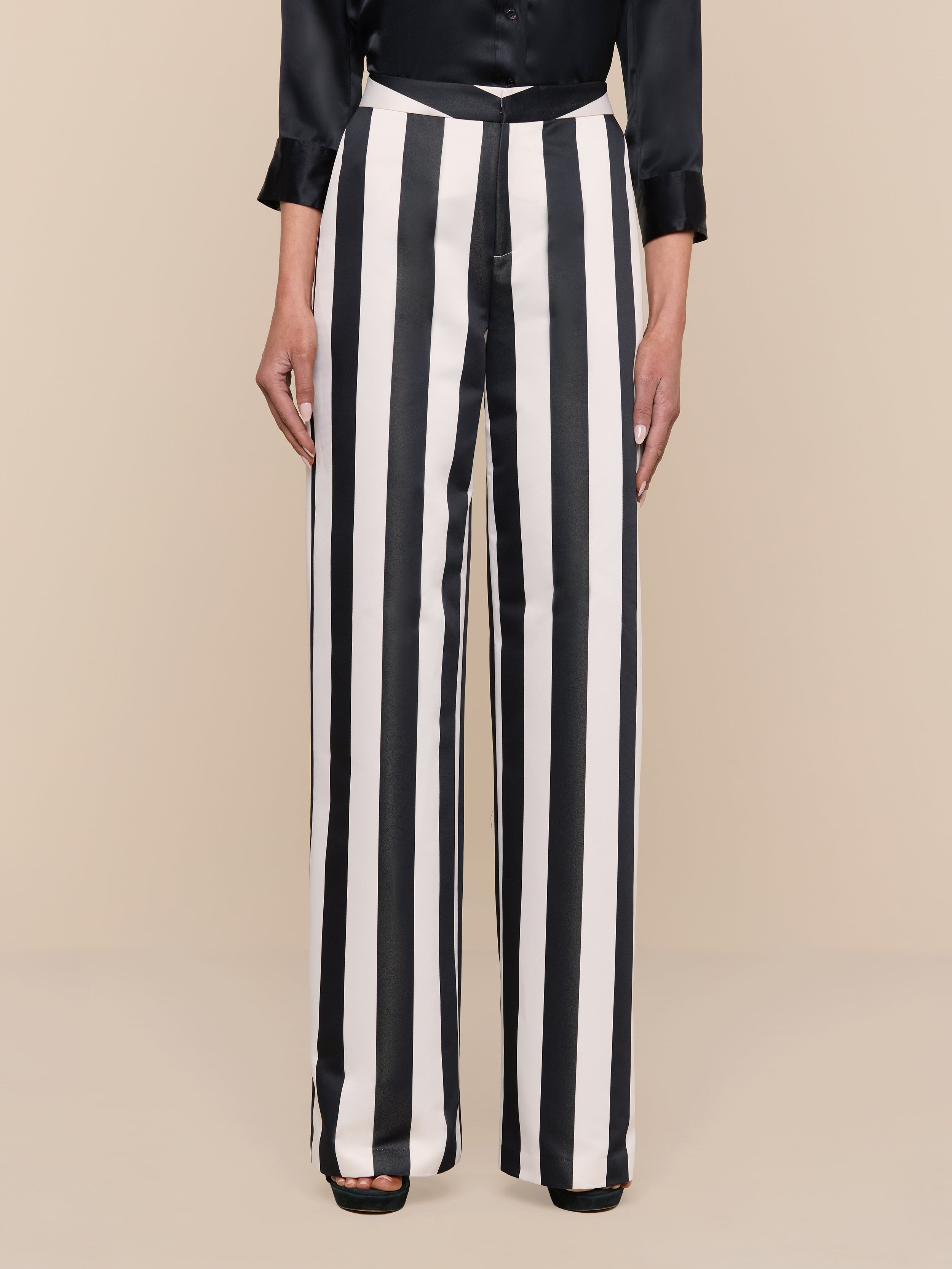 A person wearing the Livvy Satin Straight-Leg Trouser, styled in a high-rise fit with black and white stripes and wide legs, paired with a black long-sleeve top and standing against a plain white background.