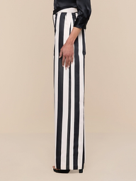 A person is shown from the shoulder down, standing sideways against a plain white background, wearing the Livvy Satin Straight-Leg Trouser in black and white stripes paired with a long-sleeve black top.