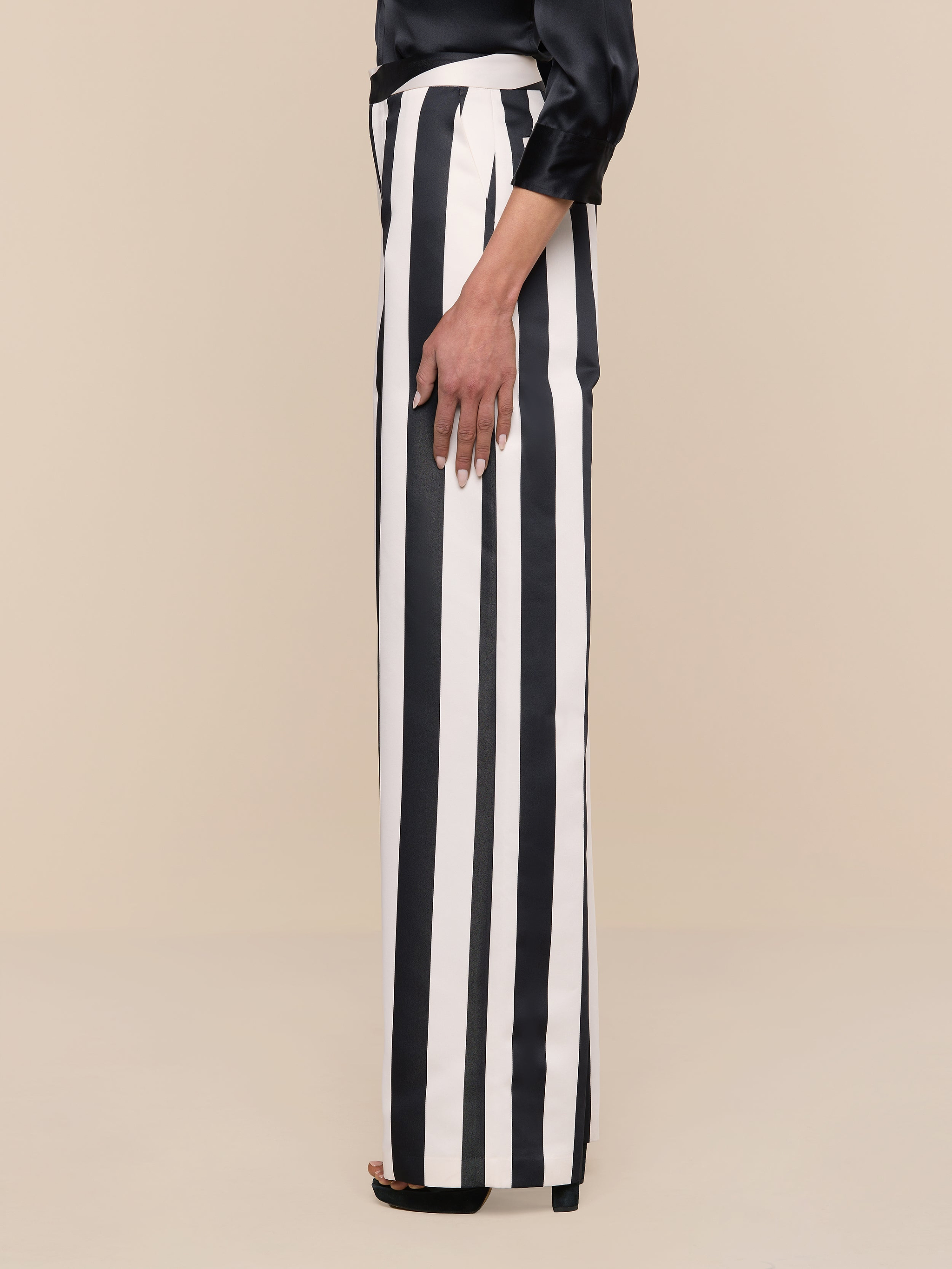 A person is shown from the shoulder down, standing sideways against a plain white background, wearing the Livvy Satin Straight-Leg Trouser in black and white stripes paired with a long-sleeve black top.