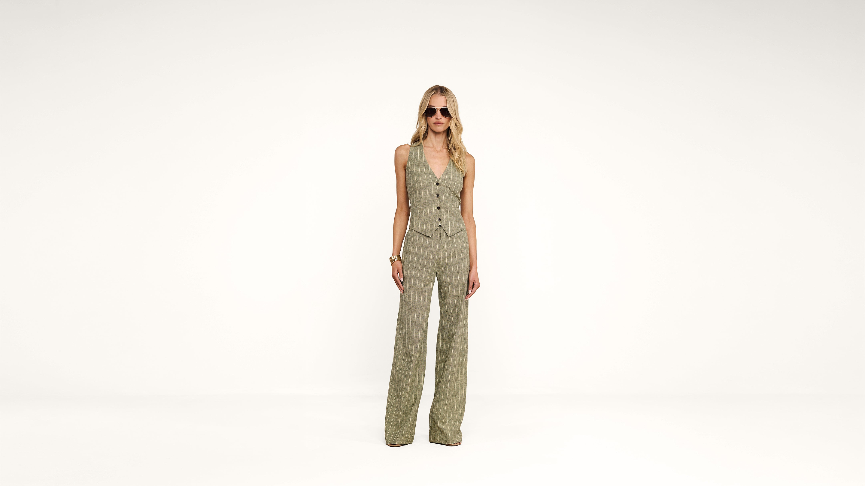 A person wearing sunglasses, a sleeveless, buttoned vest, and matching wide-leg pants stands against a plain white background.