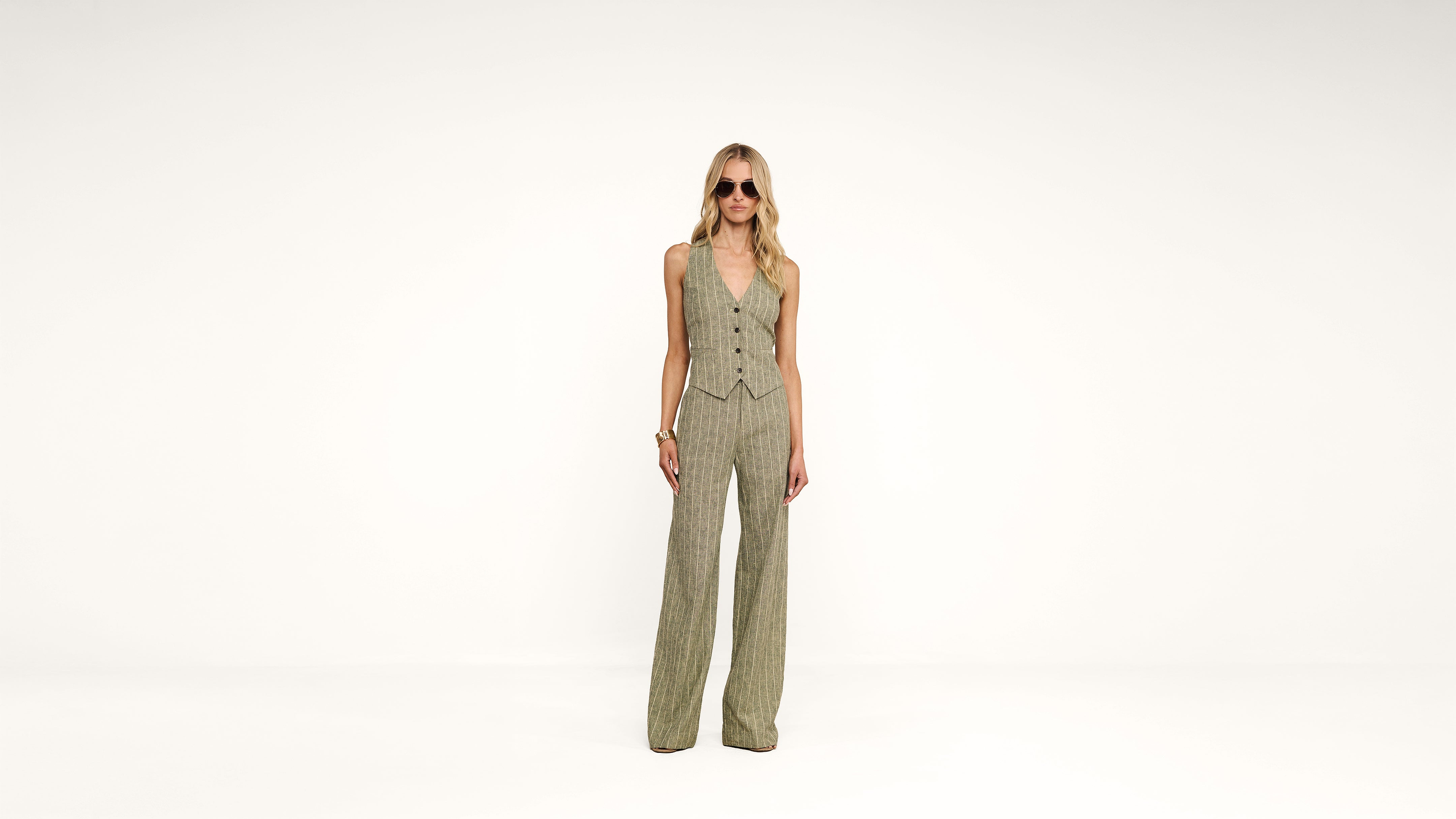 A person wearing sunglasses, a sleeveless, buttoned vest, and matching wide-leg pants stands against a plain white background.