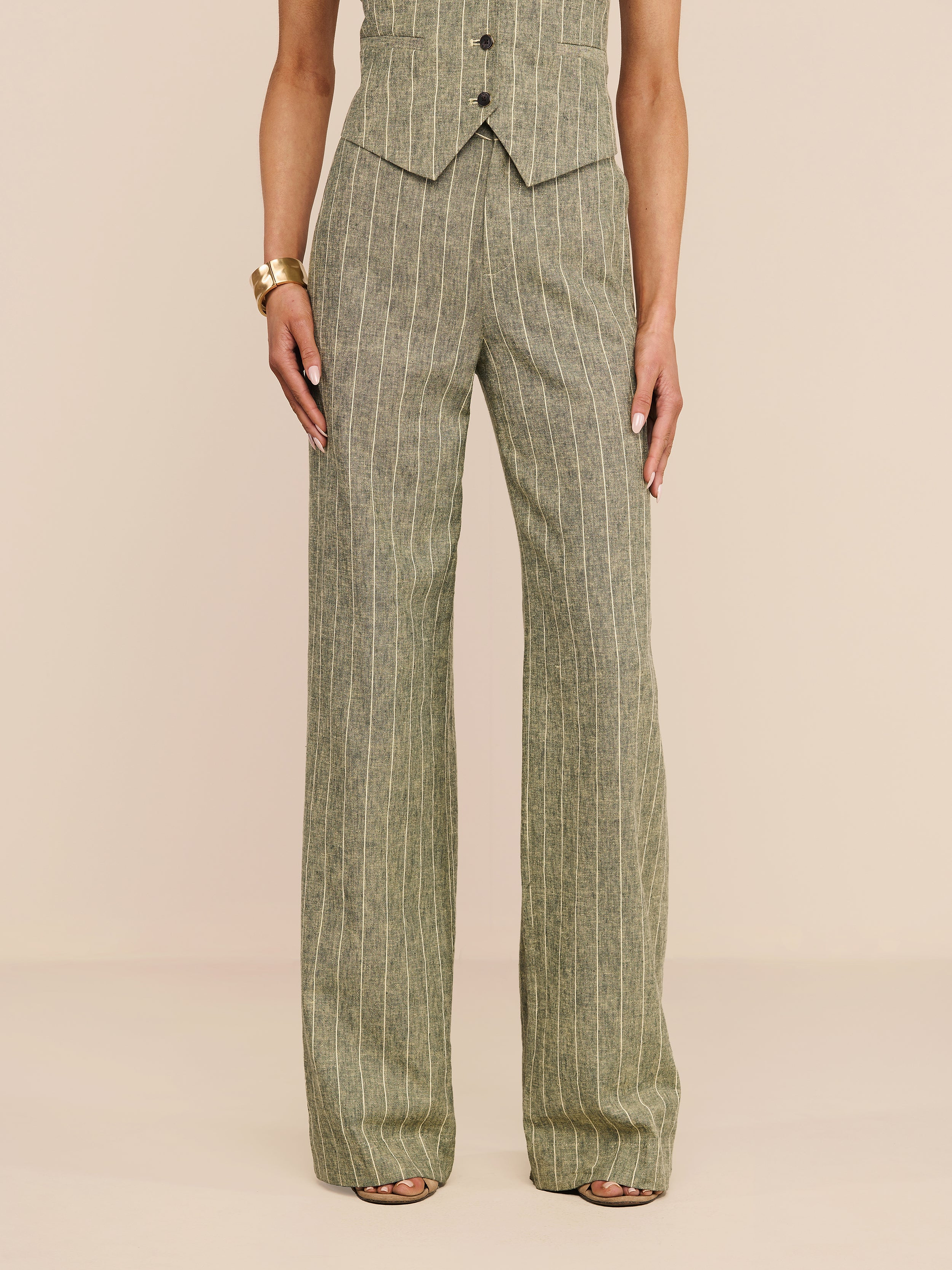A person stands against a white background, wearing the high-waisted Livvy Linen-Blend Trouser in sage green pinstripes paired with a matching vest. Their look is completed with strappy heels and a chunky gold bracelet.