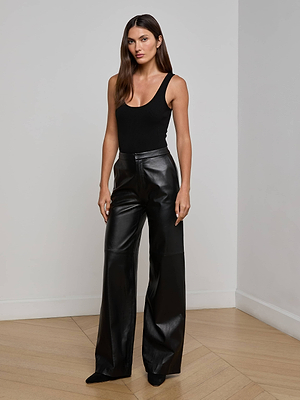 A woman with long brown hair wears the L'AGENCE Livvy Leather Trousers, a high-waisted, straight-leg style, paired with a black sleeveless top. She stands indoors on a light wood floor against a plain white wall.