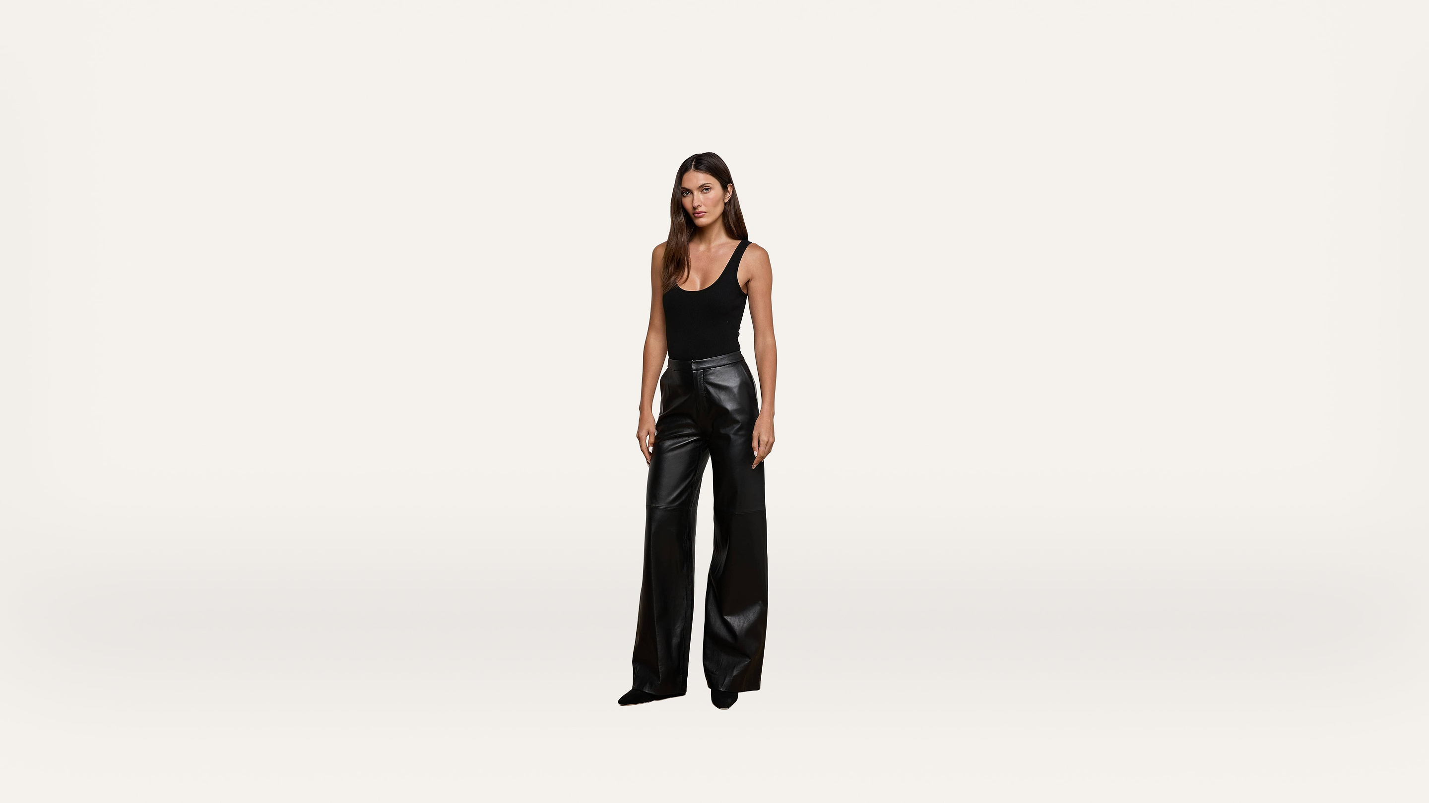 A woman stands against a plain white background, wearing a black sleeveless top and high-waisted, wide-leg black leather pants.
