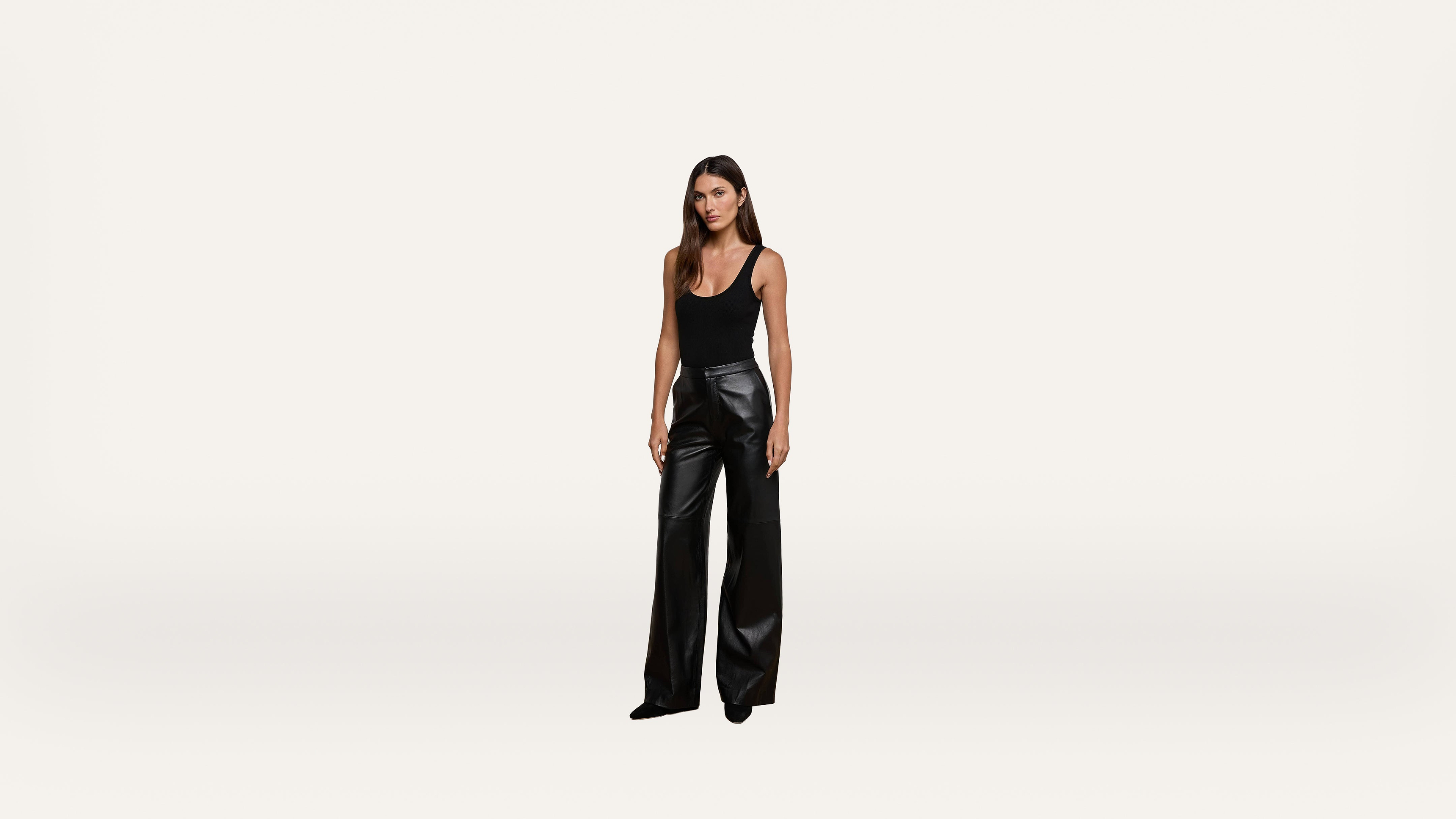 A woman stands against a plain white background, wearing a black sleeveless top and high-waisted, wide-leg black leather pants.