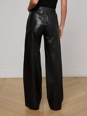 A person stands with their back to the camera, wearing L'AGENCE Livvy Leather Trousers—high-waisted, straight-leg black leather pants—and a sleeveless black top, on a light wood floor against a plain white wall.