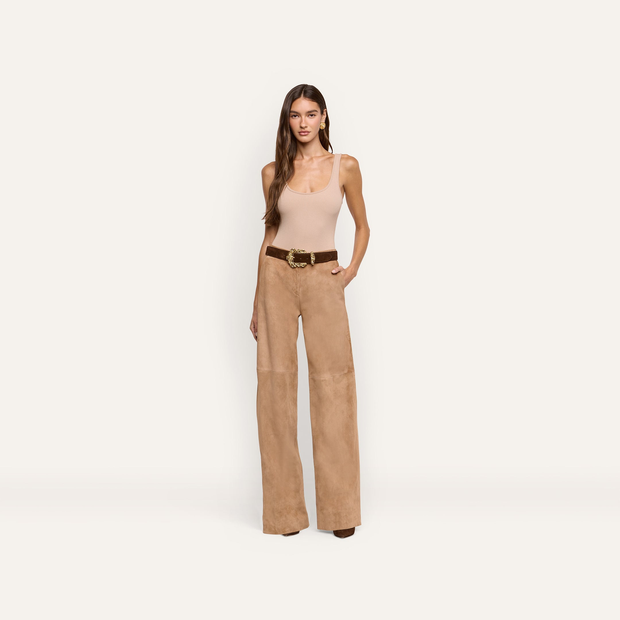 A woman wears a beige tank top and Livvy Suede Straight-Leg Trouser in light brown, secured with a brown belt featuring a gold buckle.