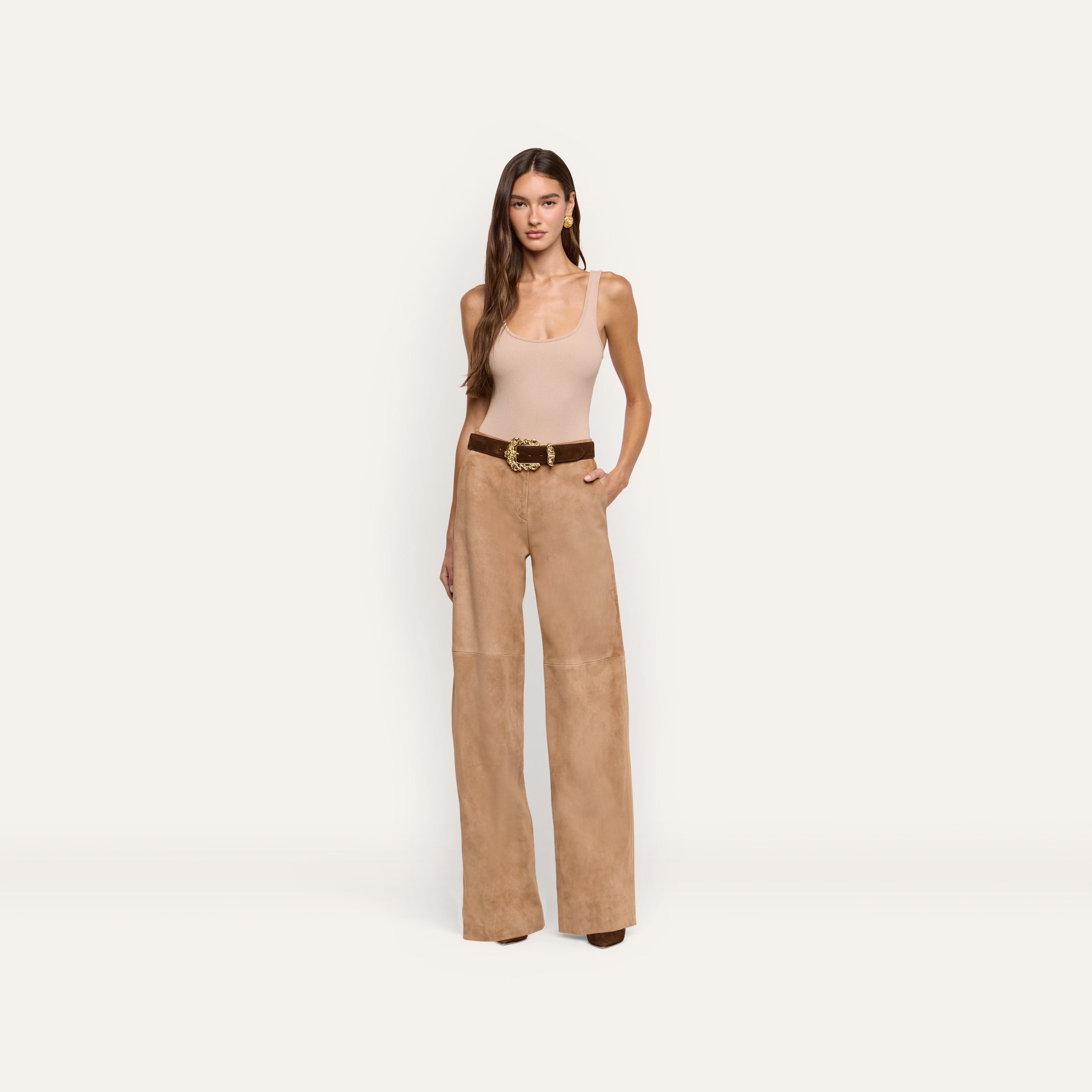 A woman wears a beige tank top and Livvy Suede Straight-Leg Trouser in light brown, secured with a brown belt featuring a gold buckle.