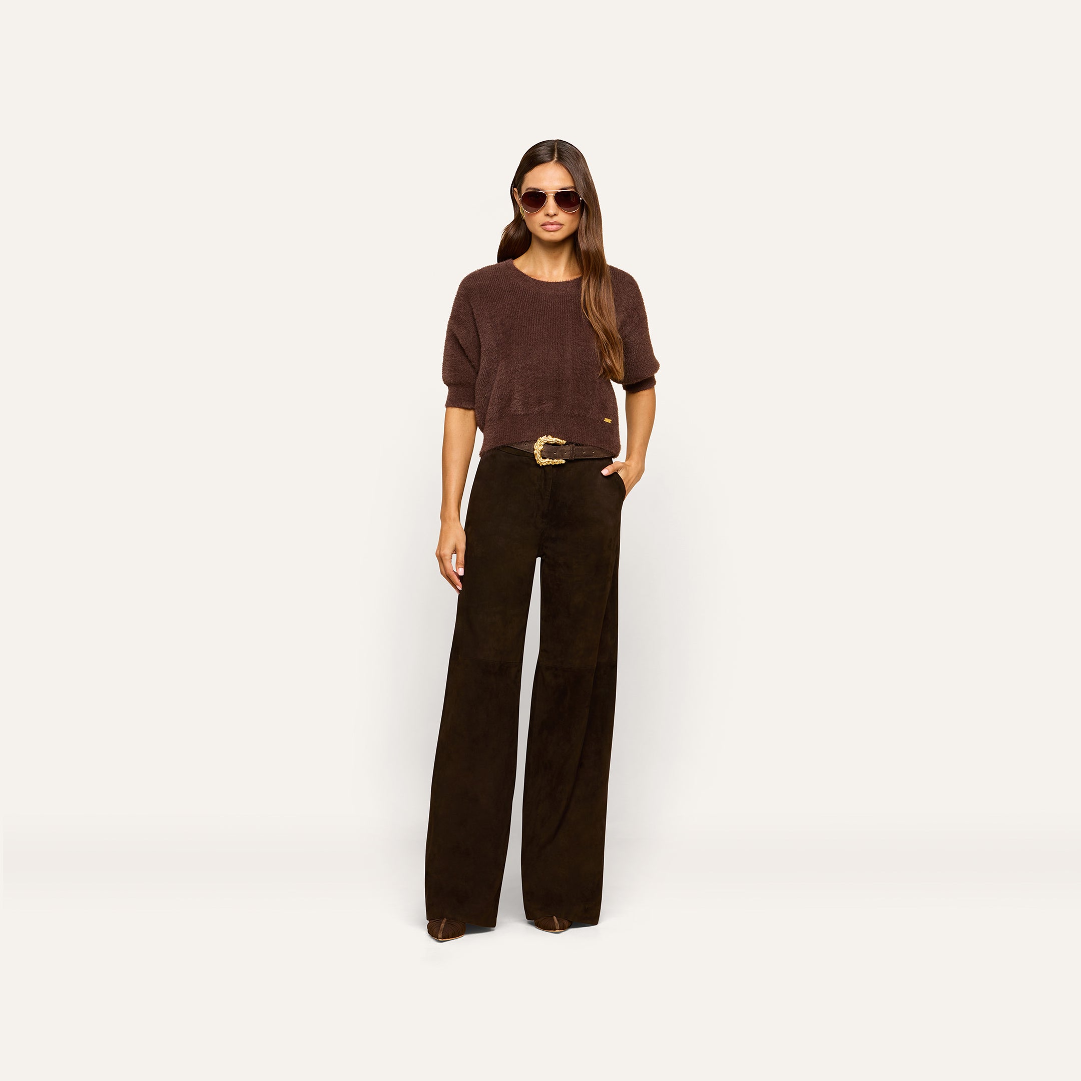 Woman wearing sunglasses, a loose brown sweater, and the Livvy Suede Trouser—a pair of high rise black wide-leg pants with a belt—standing against a plain light background with one hand in her pocket.
