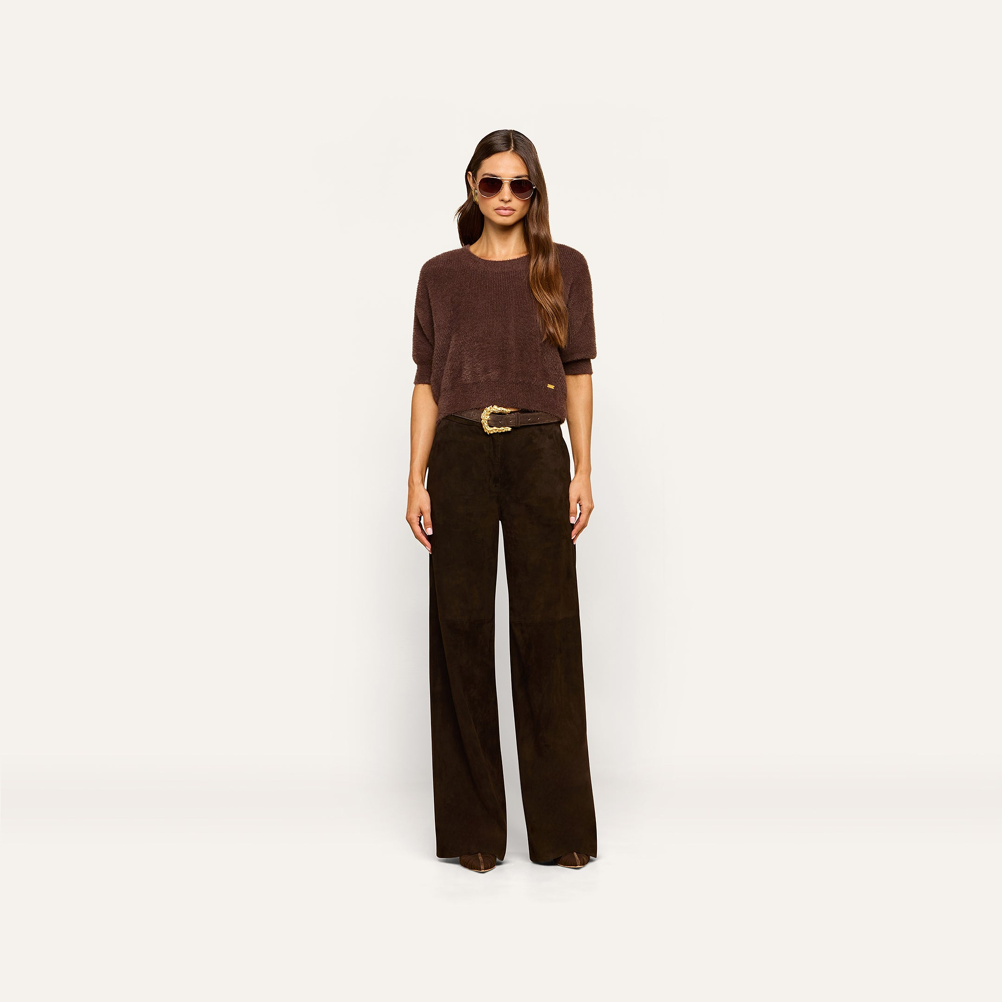 A woman wearing sunglasses, a brown short-sleeve top, and the Livvy Suede Trouser—high-rise, wide-leg dark brown pants with a large belt buckle—styled for Fall 2026 and standing against a plain light background.
