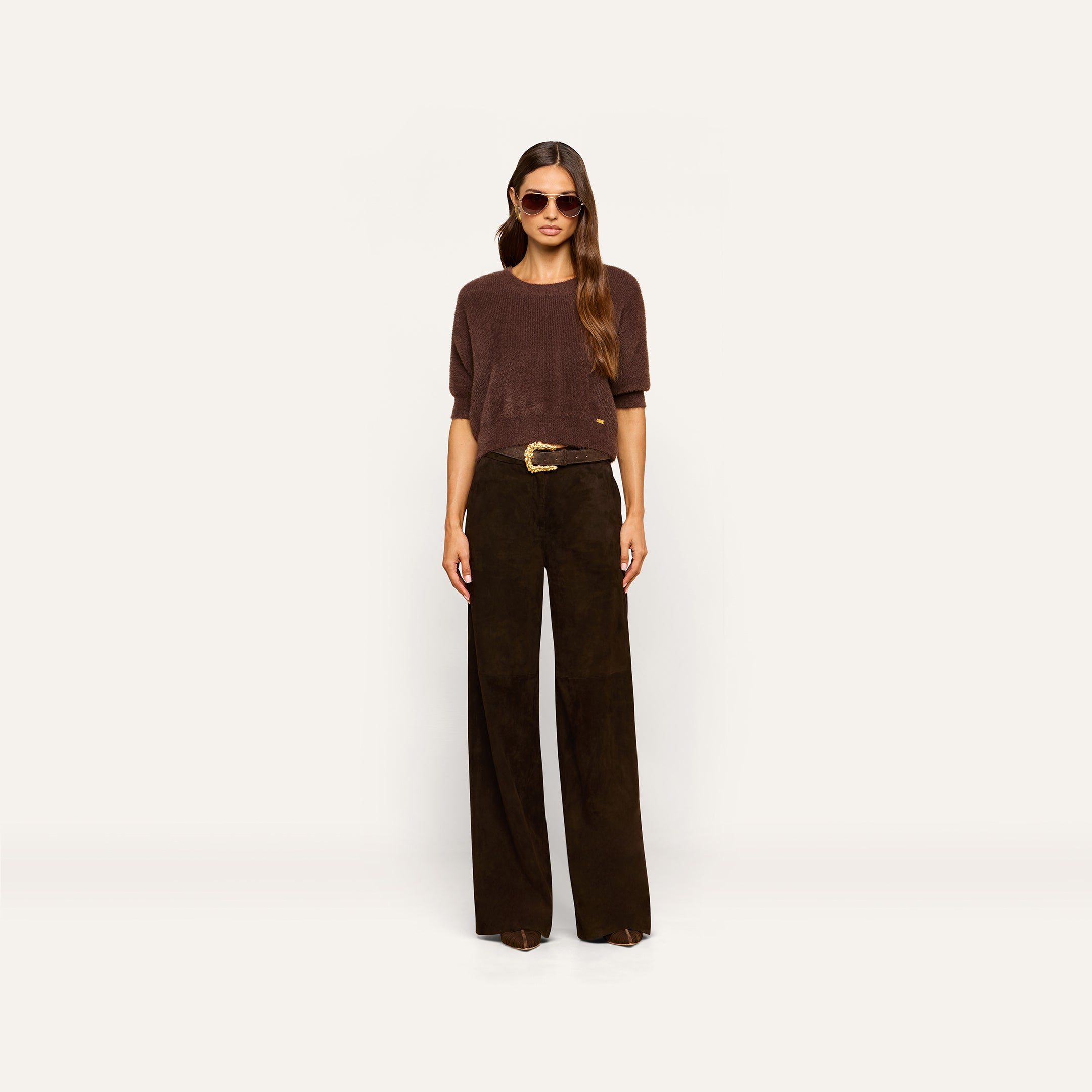 A woman wearing sunglasses, a brown short-sleeve top, and the Livvy Suede Trouser—high-rise, wide-leg dark brown pants with a large belt buckle—styled for Fall 2026 and standing against a plain light background.