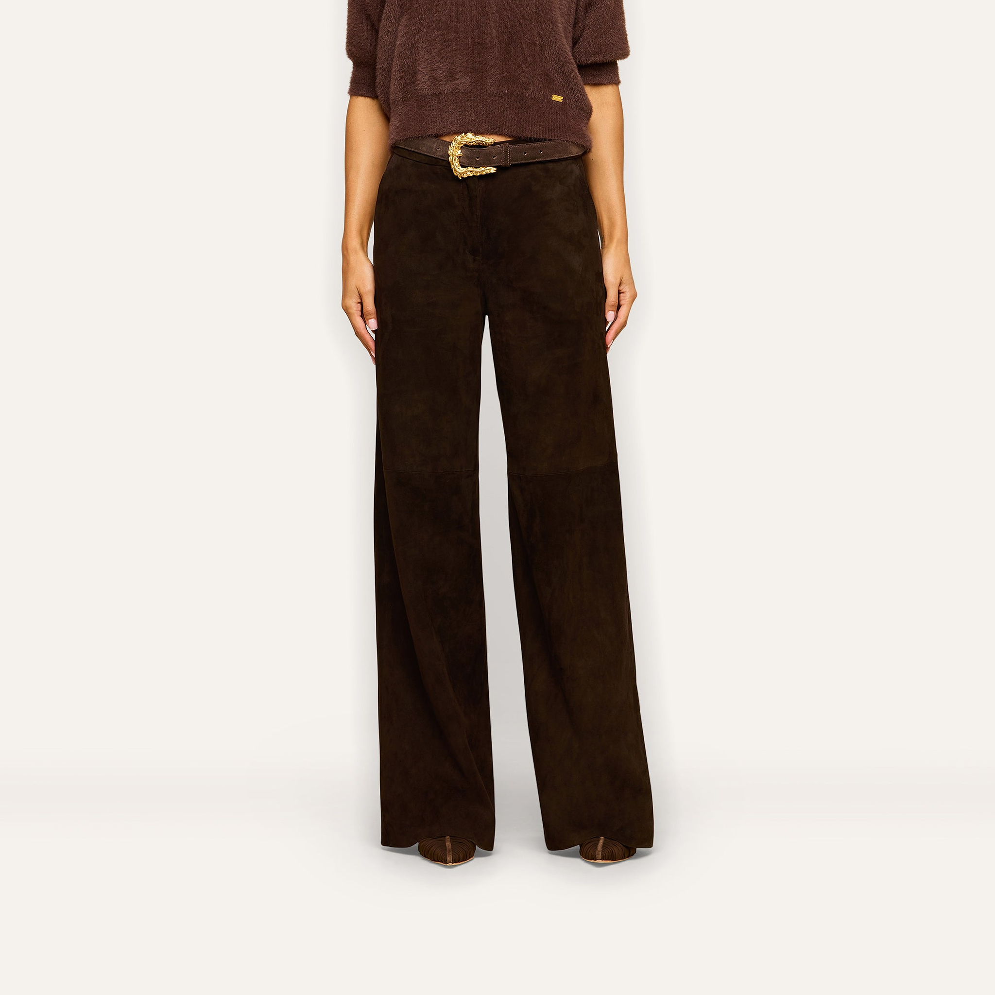 A person wears the Livvy Suede Trouser—a high-rise, straight-leg style—in suede, paired with a brown sweater tucked in and a decorative belt, standing against a plain light background. The face is not visible.