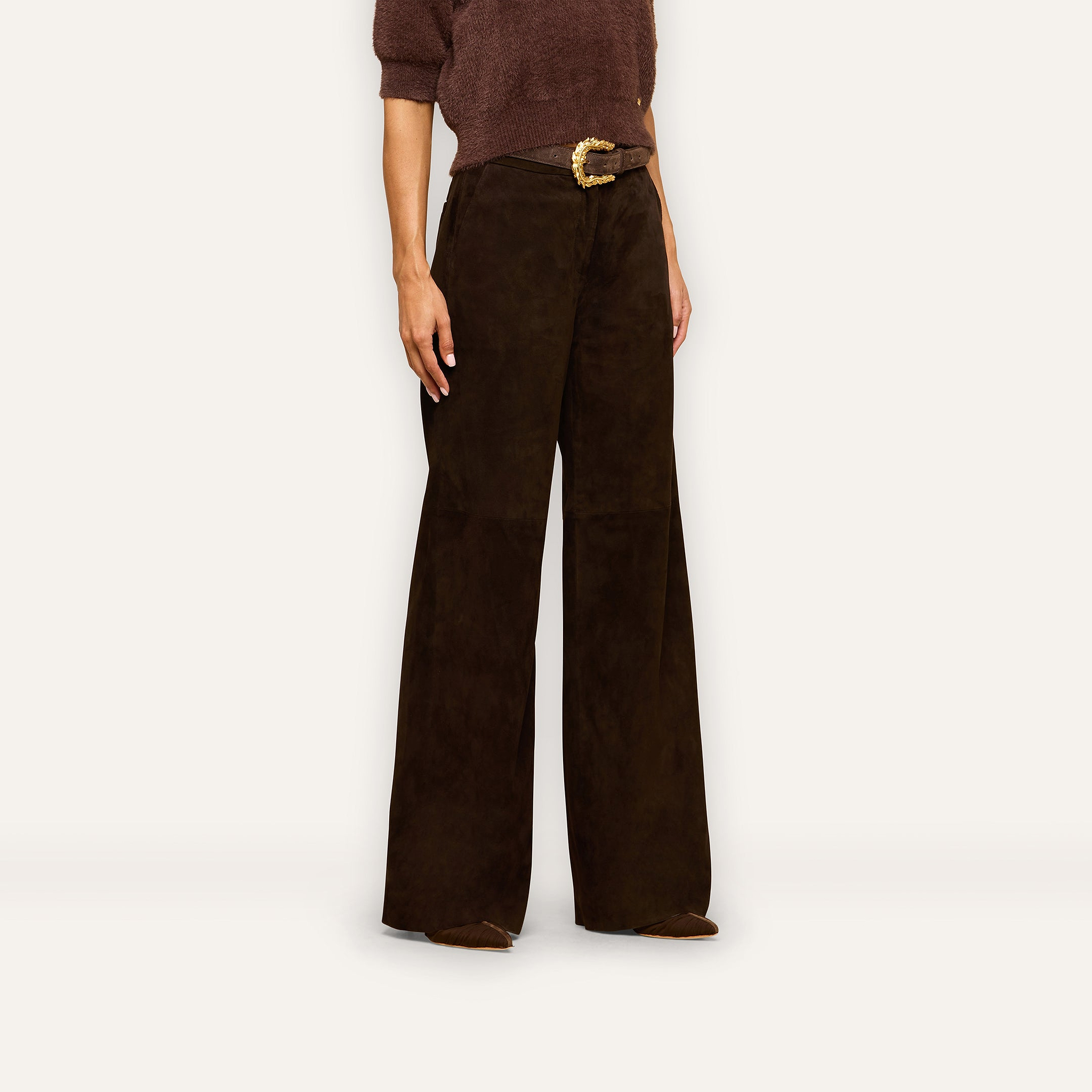 A person wearing a brown knit top, the Livvy Suede Trouser in a high-rise straight-leg silhouette, and a belt with a gold buckle stands against a plain background, capturing the essence of Fall 2026 style.