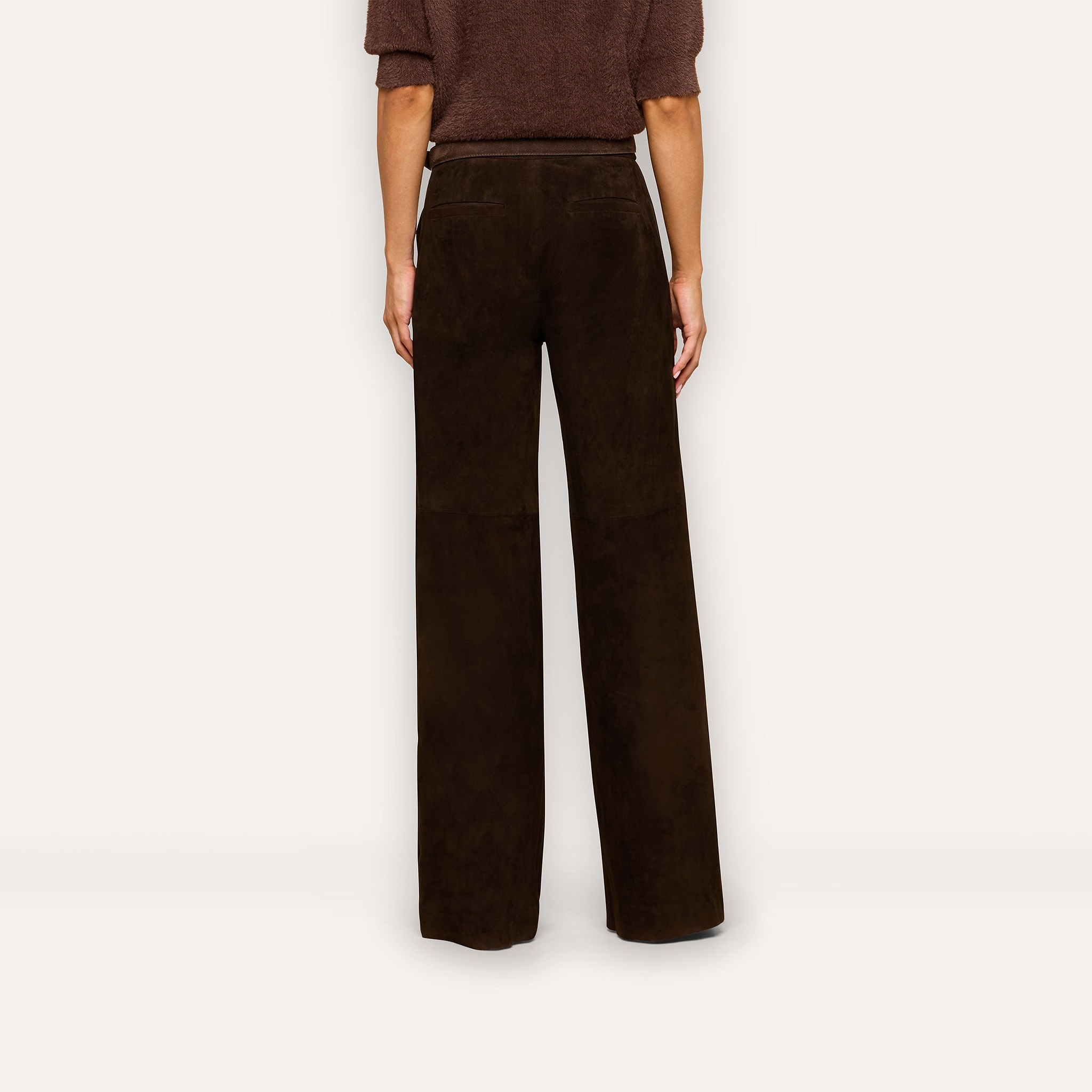 A person stands facing away, wearing the Livvy Suede Trouser—a high-rise, wide-leg pant in dark brown—paired with a short-sleeve brown top. The plain off-white background evokes a timeless Fall 2026 style.