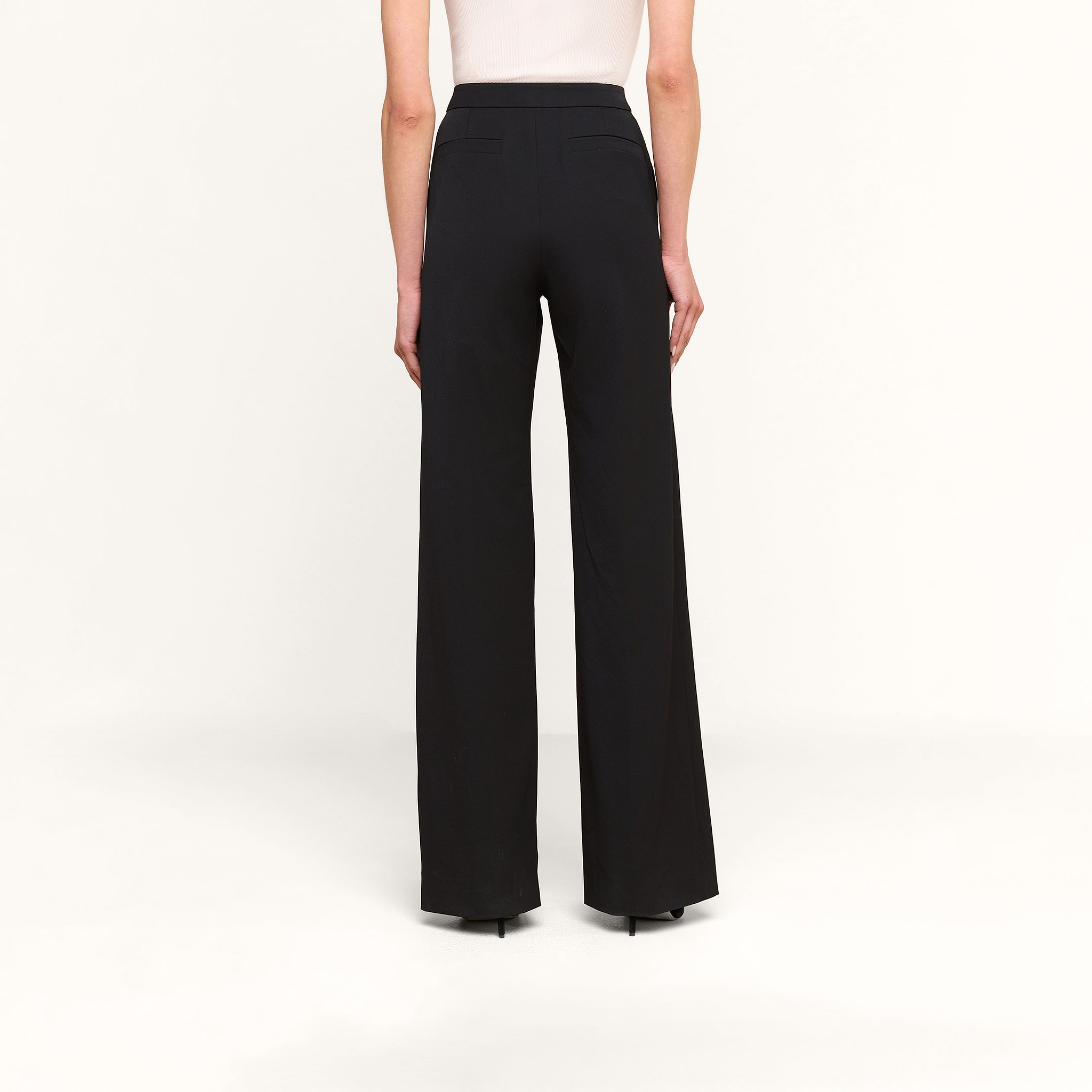 Une personne vue de dos sur un fond beige uni, portant le pantalon Livvy — un pantalon noir à taille haute et à jambes larges — associé à un haut blanc sans manches.
