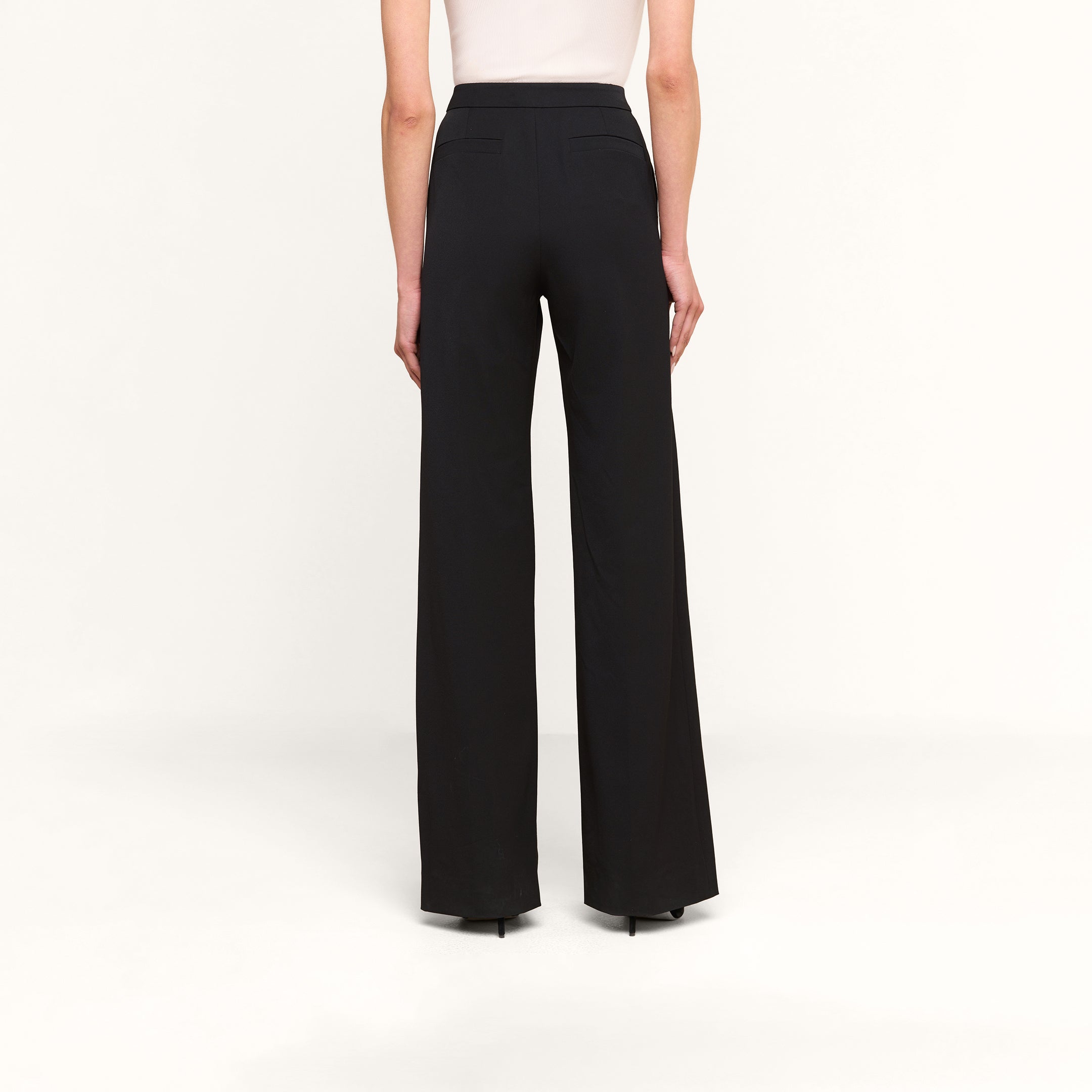 Une personne vue de dos sur un fond beige uni, portant le pantalon Livvy — un pantalon noir à taille haute et à jambes larges — associé à un haut blanc sans manches.
