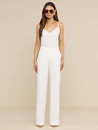 A woman stands against a plain white background, wearing the Livvy Trouser—a high-rise, wide-leg pant styled with a sleeveless white jumpsuit top. She completes the look with nude heels and large round sunglasses, her long hair worn loose and her hands by her sides.