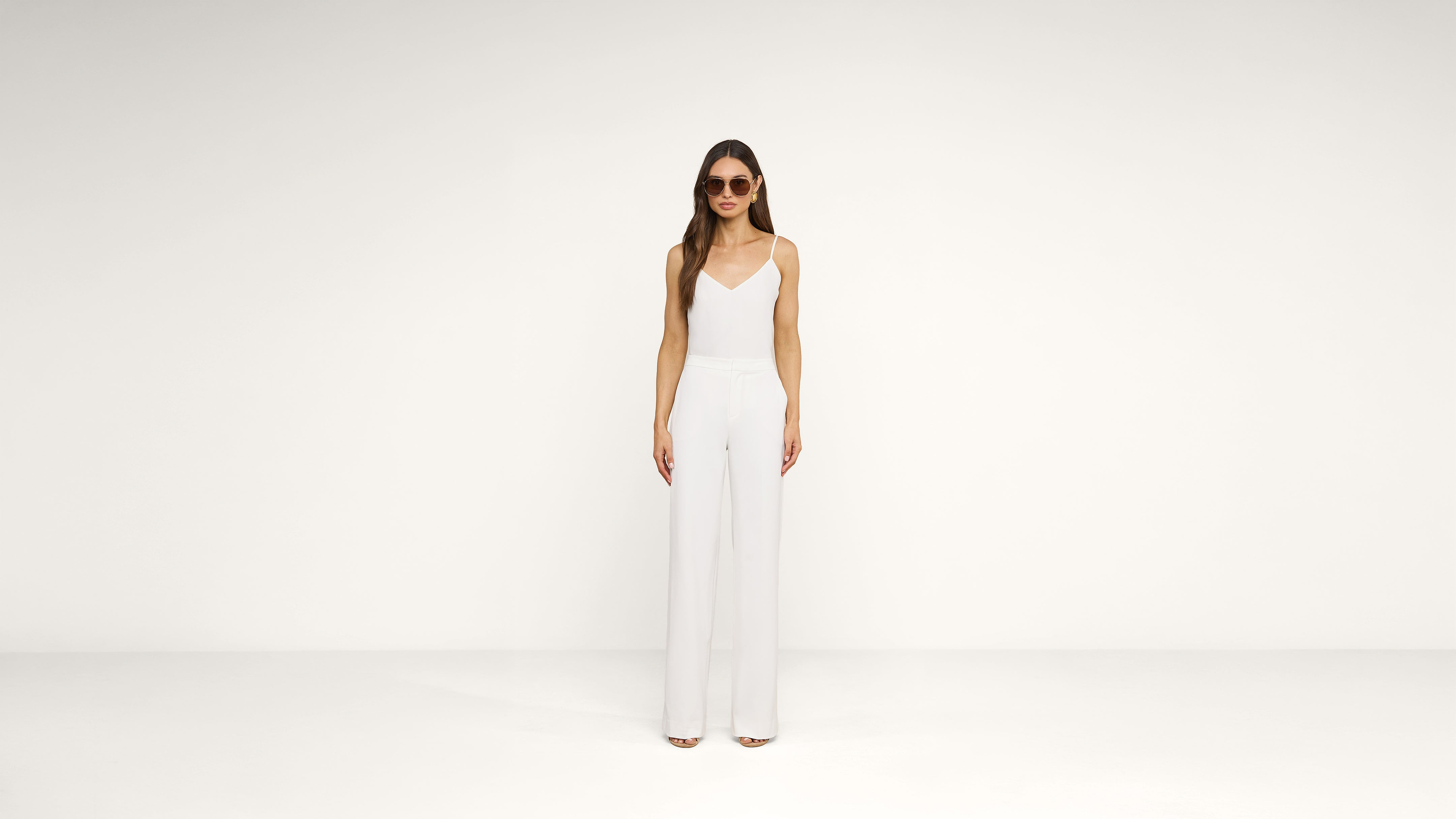 A woman wearing sunglasses, a sleeveless white top, and white wide-leg pants stands against a plain white background.