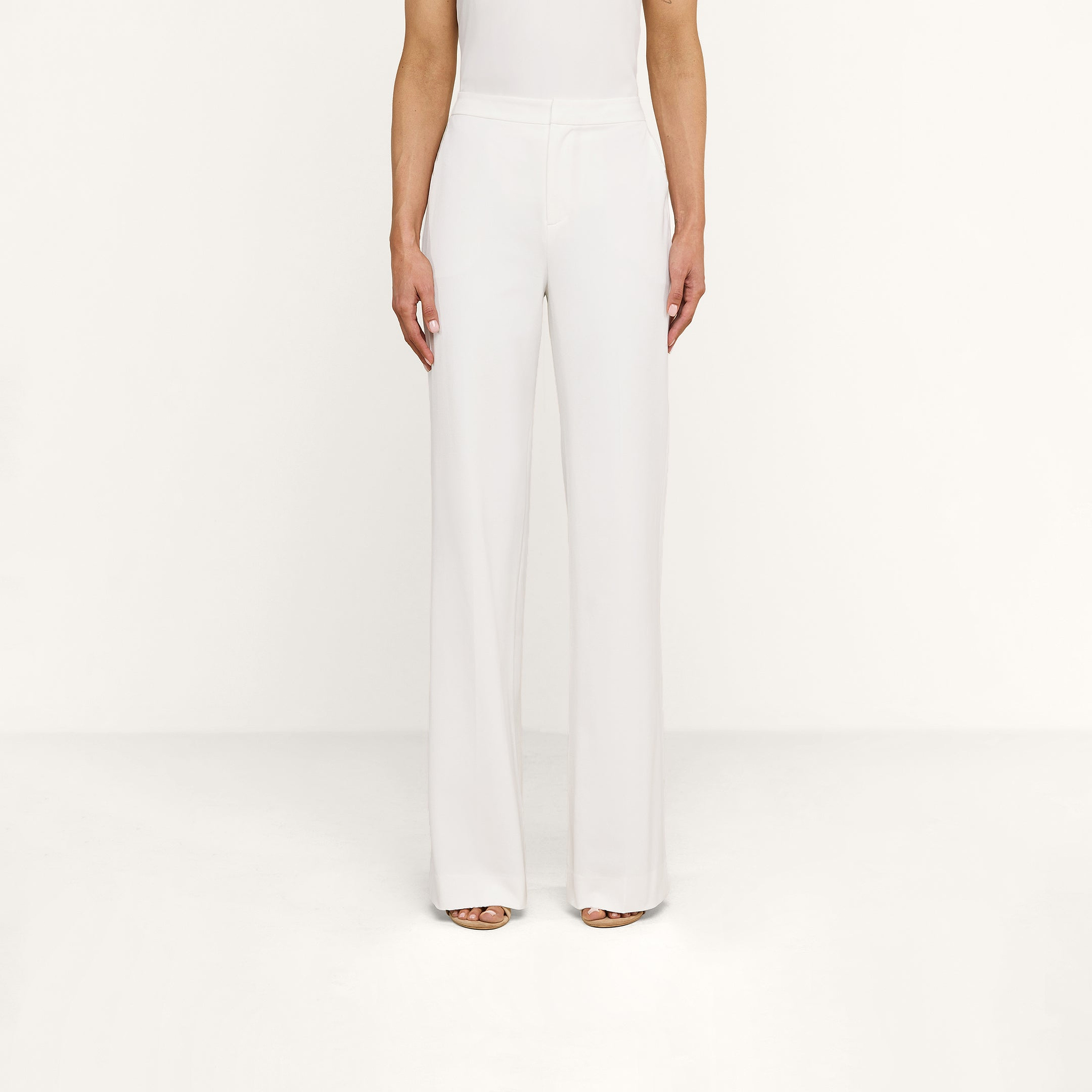 A person wearing the Livvy Trouser, a pair of high-rise, wide-leg white trousers, and open-toe heels stands against a plain white background with their arms relaxed by their sides.