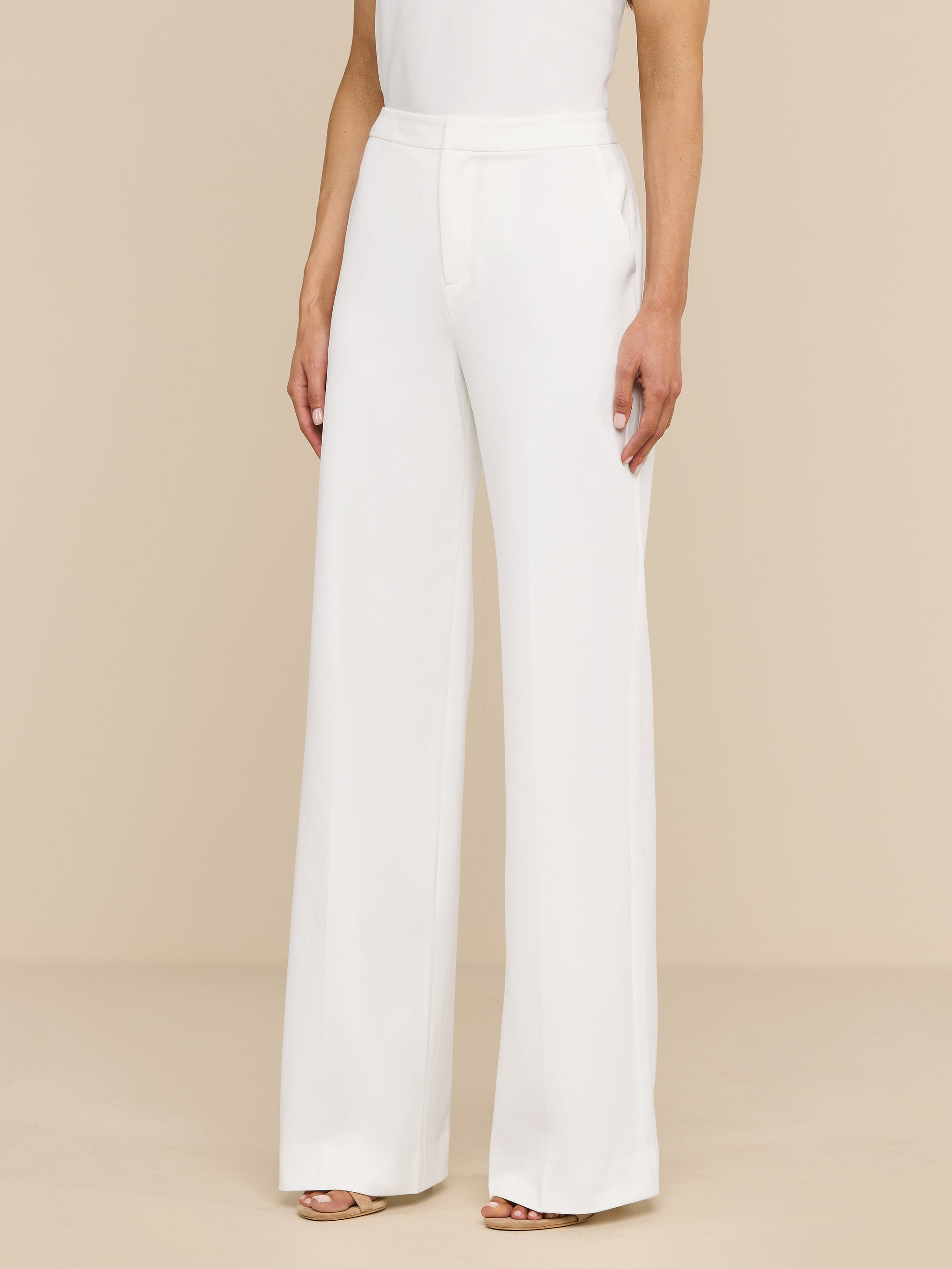 A woman stands against a plain white background, wearing the high rise, wide leg Livvy Trouser and a sleeveless white top. Only her body from the shoulders down is visible.
