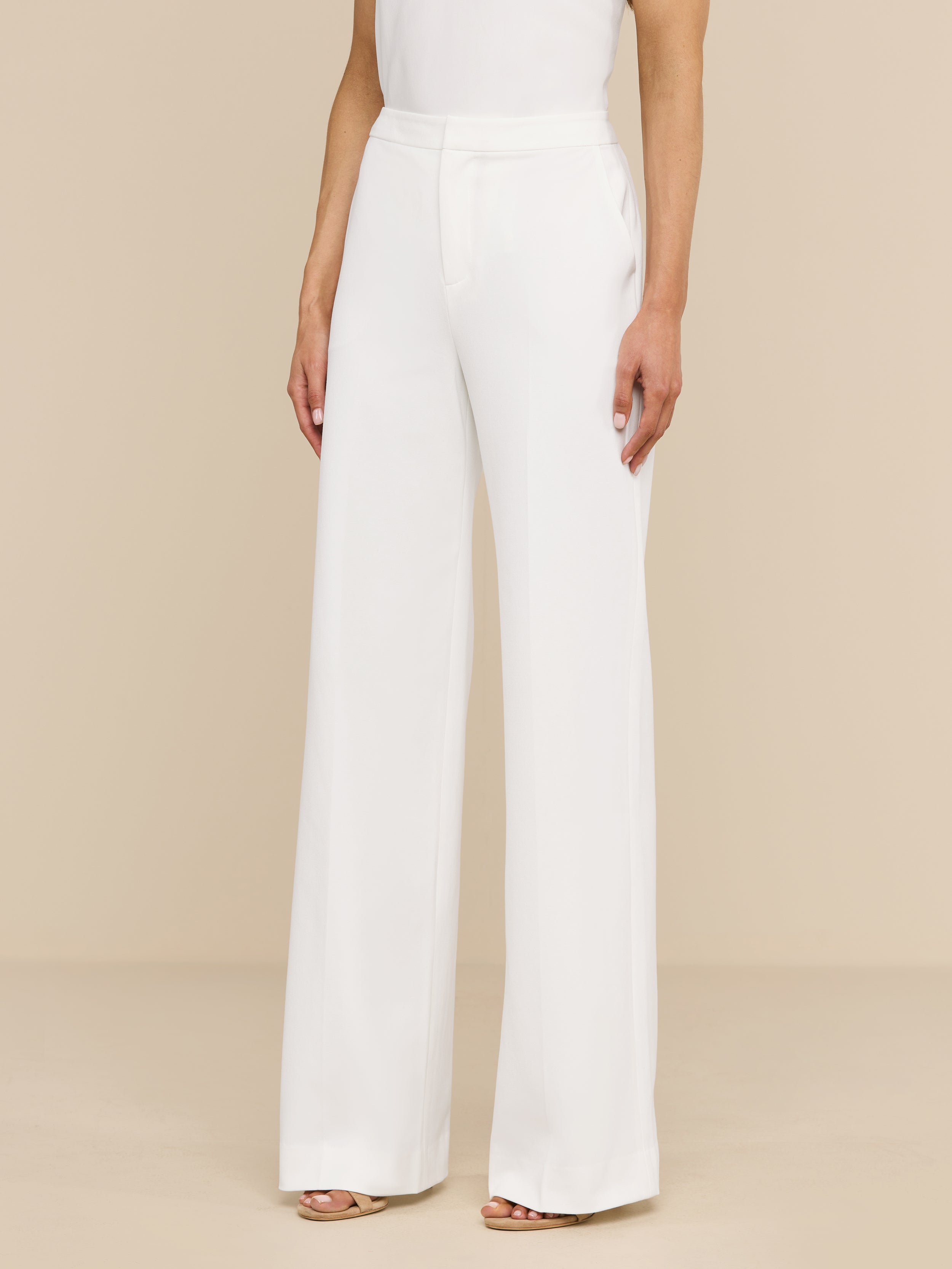 A woman stands against a plain white background, wearing the high rise, wide leg Livvy Trouser and a sleeveless white top. Only her body from the shoulders down is visible.