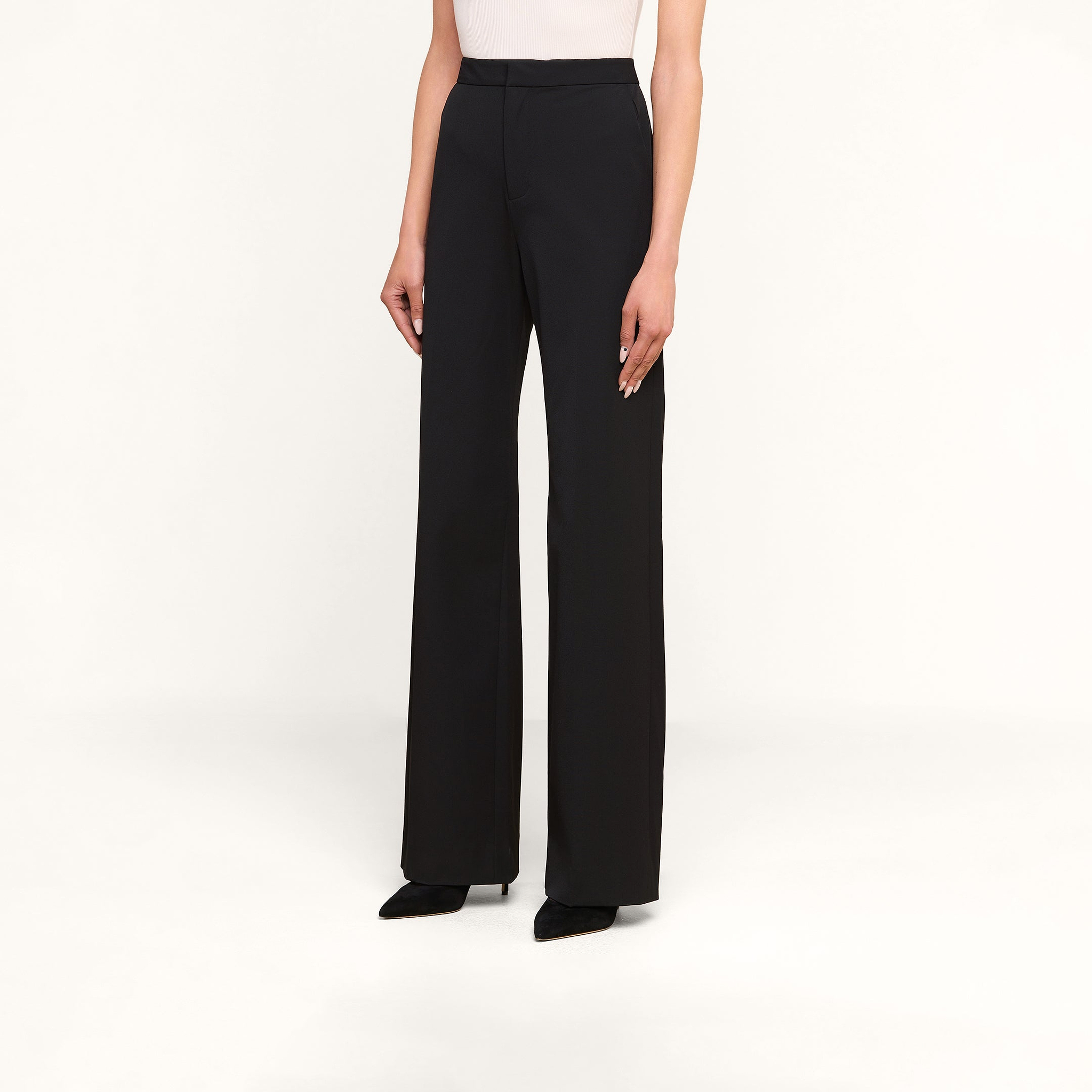 Wearing the Livvy Trouser in black—a high-waisted, wide-leg style—paired with a white top and black pointed-toe shoes, this look is set against a plain beige background, making it ideal for transitioning from the office to dinner.