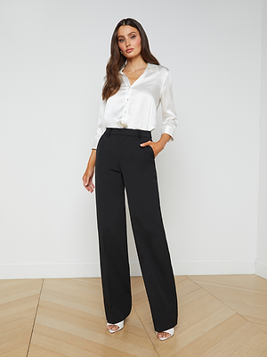 A woman stands indoors against a white wall, wearing L'AGENCE Livvy Trousers and a white satin blouse with open-toe heels. One hand in her pocket, she exudes confidence—the perfect office-to-dinner look.