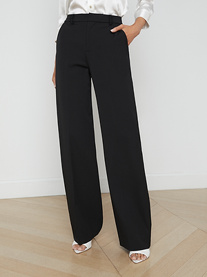 A person wearing the L'AGENCE Livvy Trouser, a black wide-leg pant, and a white satin blouse stands on light wood flooring with one hand in their pocket and white open-toe heels—ideal for office-to-dinner style.