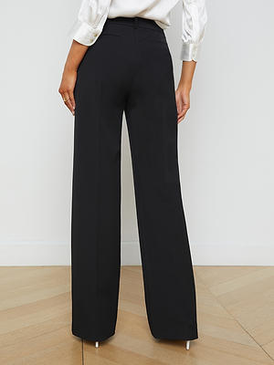 A person in the L'AGENCE Livvy Trouser—high-waisted, wide-leg black pants—and a white satin blouse stands on a light wooden floor, facing away for a refined office-to-dinner look.