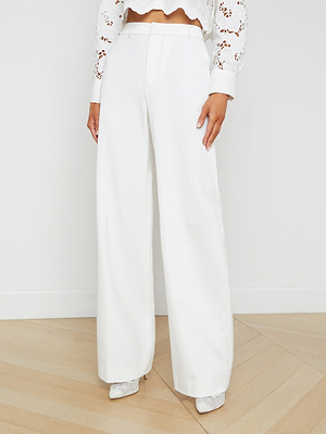 A woman wears the Livvy Trouser, a high-rise, wide-leg white trouser, paired with a white long-sleeve lace crop top and pointed-toe shoes. She stands on a light wood floor against a plain white wall.