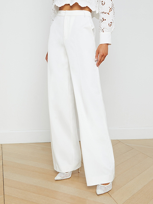 A woman wears the Livvy Trouser, a high-rise wide leg pant in white, styled with a white lace crop top and pointed-toe heels, standing on light wood herringbone flooring against a plain white wall.