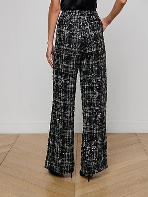 A person stands on a light wooden floor facing away from the camera, wearing L'AGENCE Livvy Tweed Trouser—high-waisted, black and white sequined tweed pants—paired with black heels against a plain white wall.