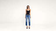A woman wearing sunglasses, a black tank top, blue ripped jeans, a black belt, and black heels stands against a plain white background.