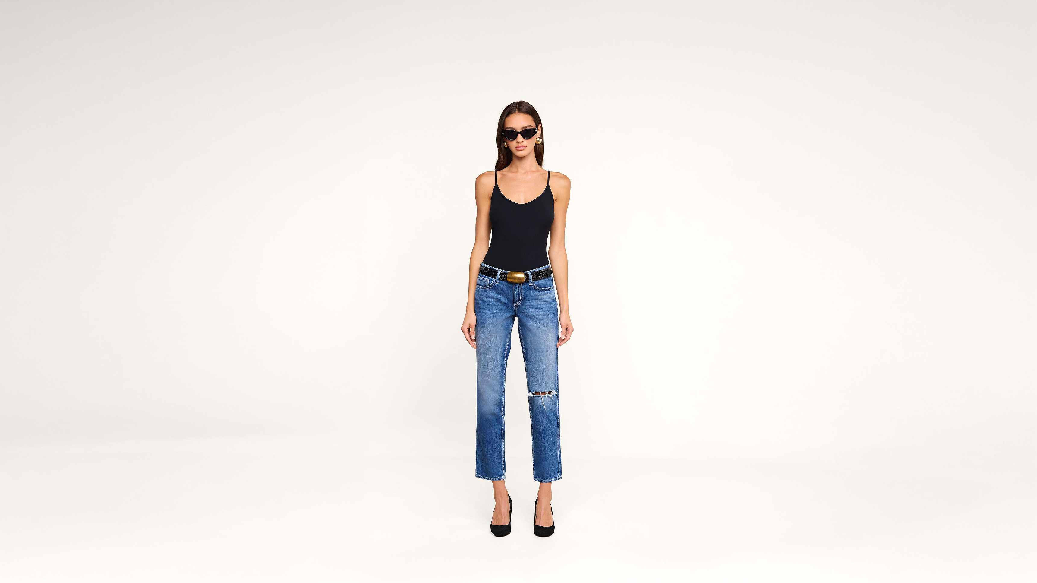 A woman wearing sunglasses, a black tank top, blue ripped jeans, a black belt, and black heels stands against a plain white background.