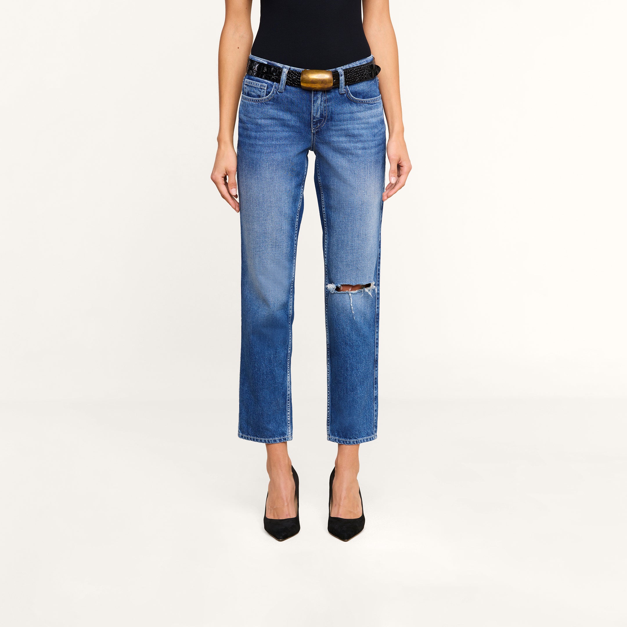 Wearing a black sleeveless top, a pair of Nevia Slouchy Straight-Leg Jeans in vintage-inspired blue all-cotton denim with a rip on one knee, a black belt with a gold buckle, and black heels, the person stands against a plain light background.