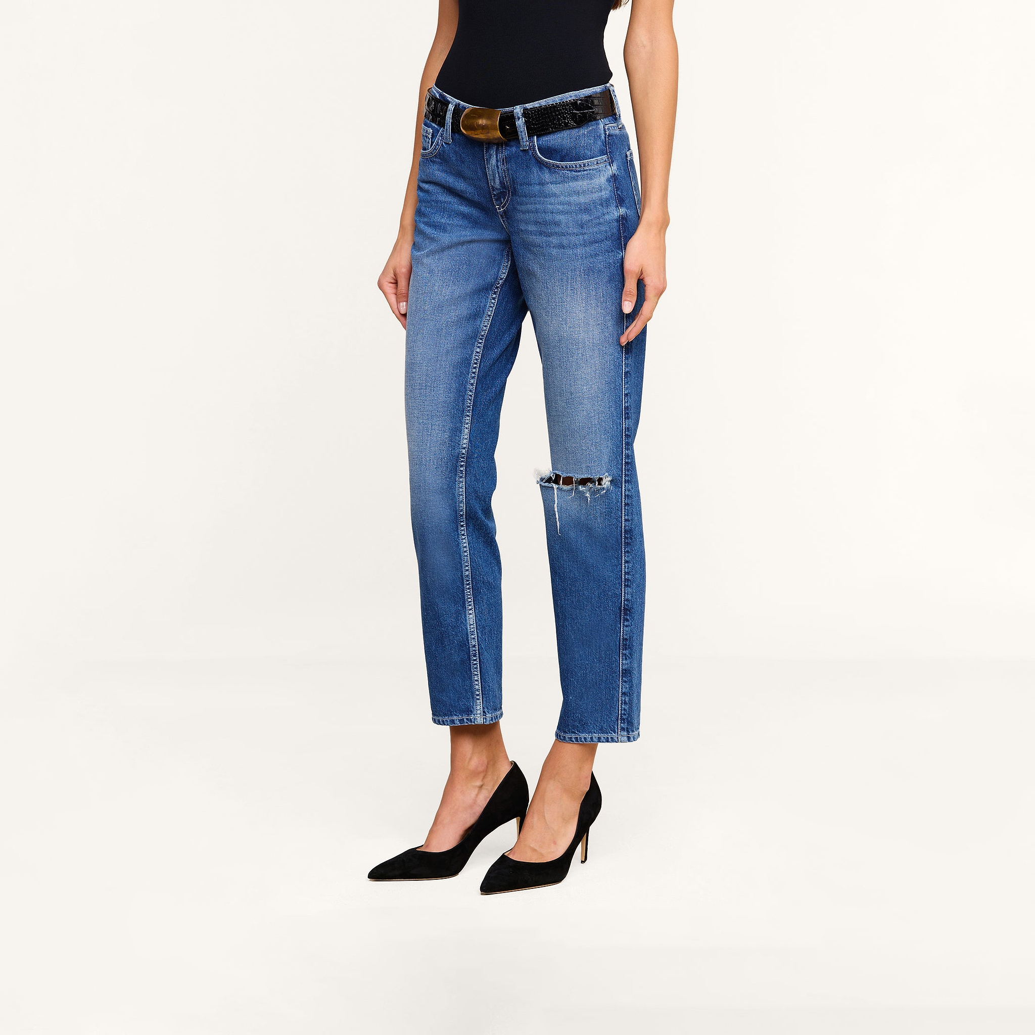 Woman wearing Nevia Slouchy Straight-Leg Jean, featuring vintage-inspired all-cotton denim in blue with a ripped knee, paired with a black belt, black sleeveless top, and black high heels, standing against a plain white background.
