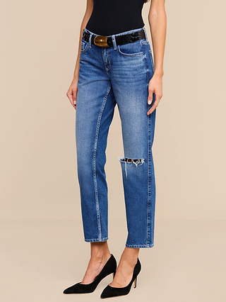 Wearing the Nevia Slouchy Straight-Leg Jean in vintage-inspired blue denim with a ripped knee, styled with a black belt, black sleeveless top, and black high heels, this woman stands against a beige background.