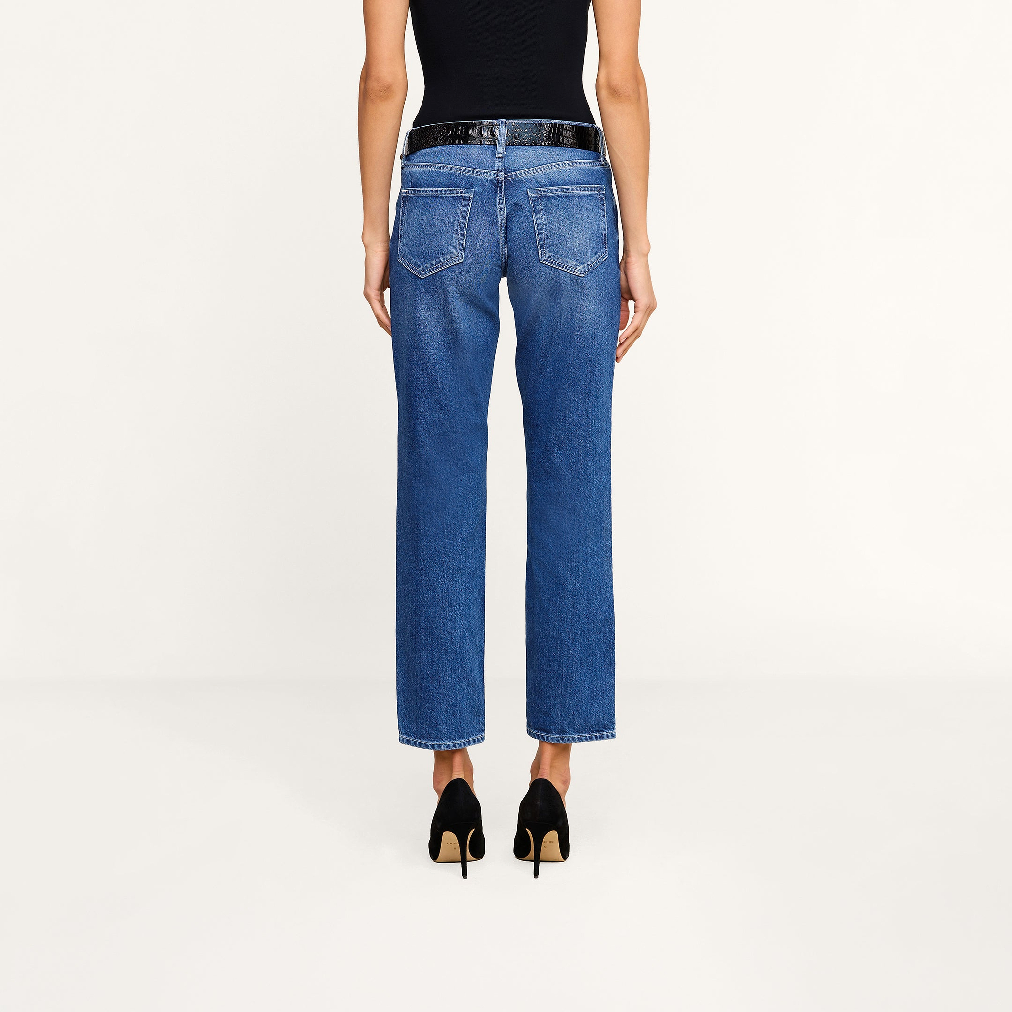 A person stands facing away, wearing a black sleeveless top, the Nevia Slouchy Straight-Leg Jean in vintage-inspired blue high-waisted all-cotton denim, and black high-heeled shoes against a plain white background.