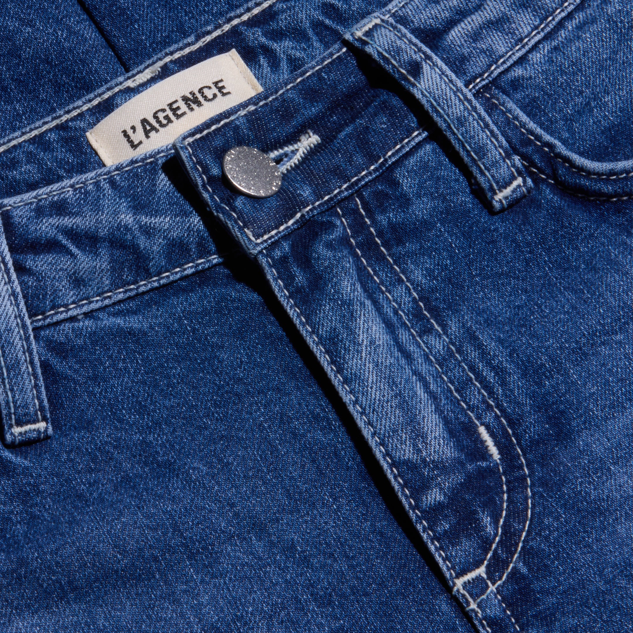 Close-up of the Nevia Slouchy Straight-Leg Jean in blue denim, showcasing a visible metal button, belt loops, and LAGENCE brand label on the waistband. Crafted from all-cotton denim with a classic textured pattern and contrast stitching, these jeans offer a vintage-inspired look.