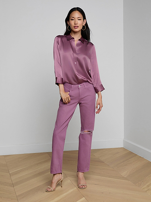 A woman stands on a wooden floor indoors, wearing a shiny mauve button-up blouse, Nevia Slouchy Straight-Leg Jean with knee rip, and nude high-heeled sandals. She poses with one hand in her pocket against a plain white wall.