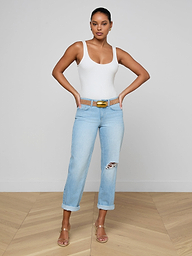 A woman poses with hands on hips against a plain white wall, wearing a white tank top and Nevia Slouchy Straight-Leg Jean in light blue denim, styled with a brown belt and beige heeled sandals on a wooden floor.