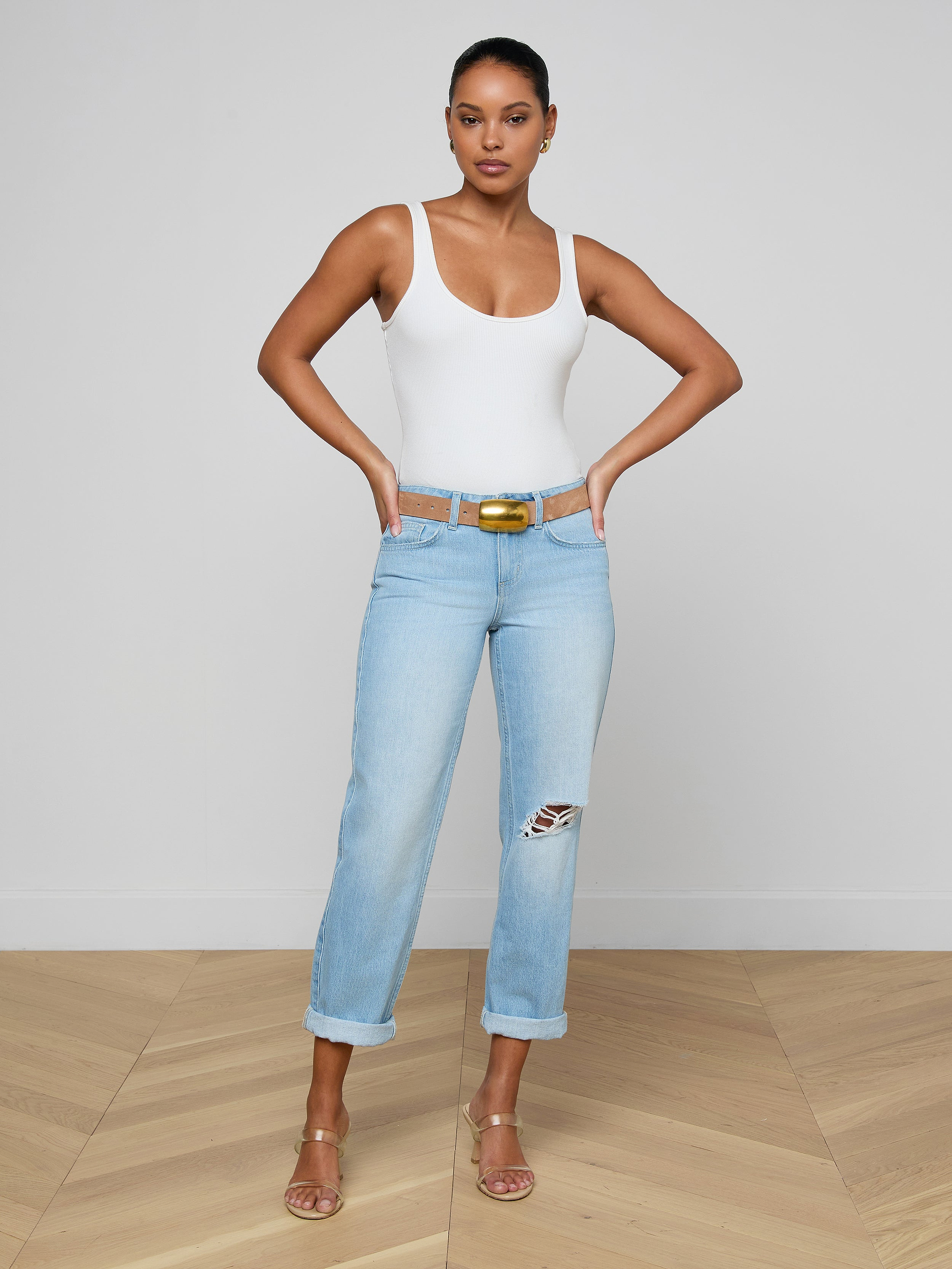 A woman poses with hands on hips against a plain white wall, wearing a white tank top and Nevia Slouchy Straight-Leg Jean in light blue denim, styled with a brown belt and beige heeled sandals on a wooden floor.