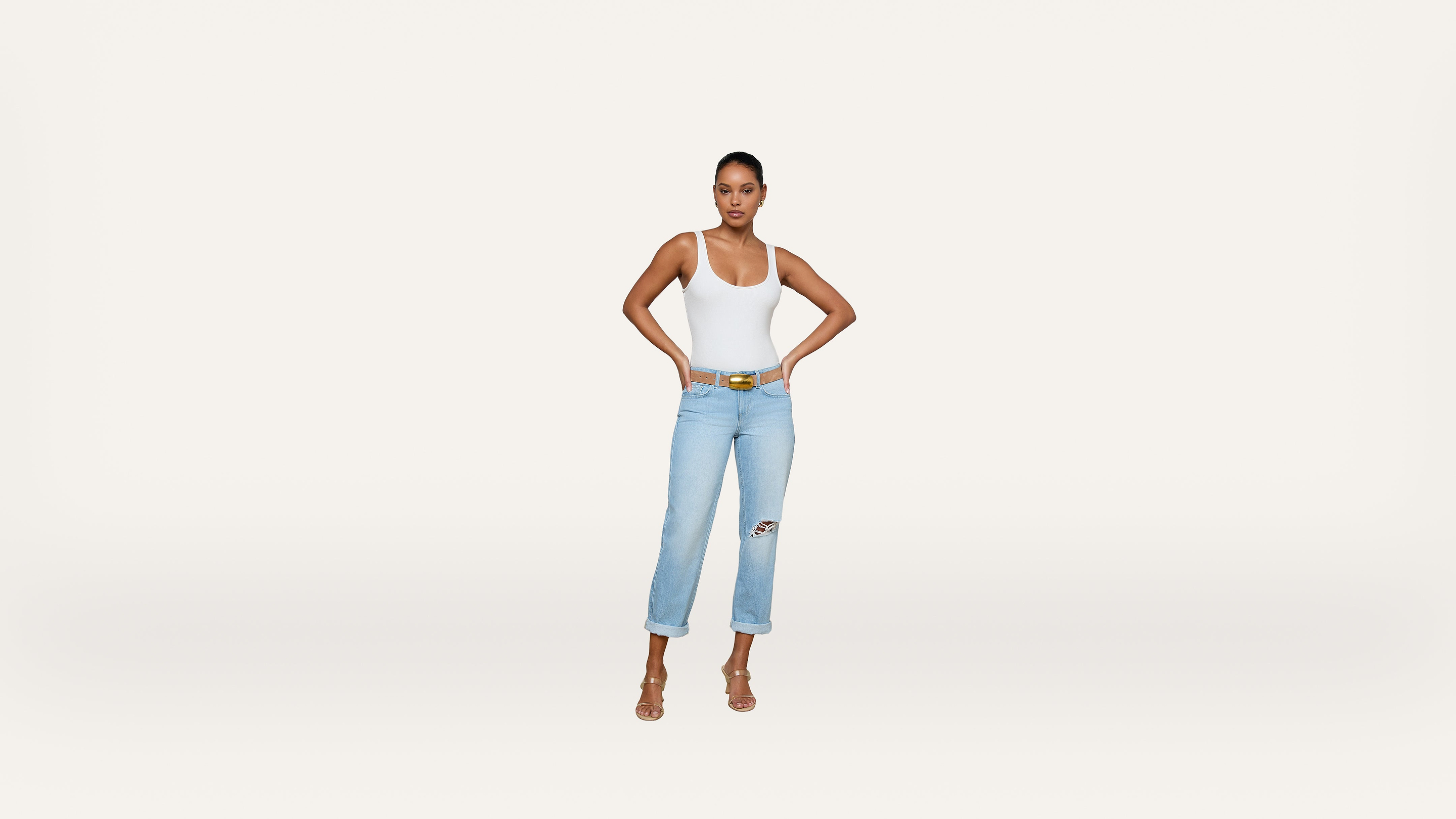 A woman stands against a plain white background, wearing a white tank top, light blue ripped jeans, a yellow belt, and heeled sandals, with her hands on her hips.