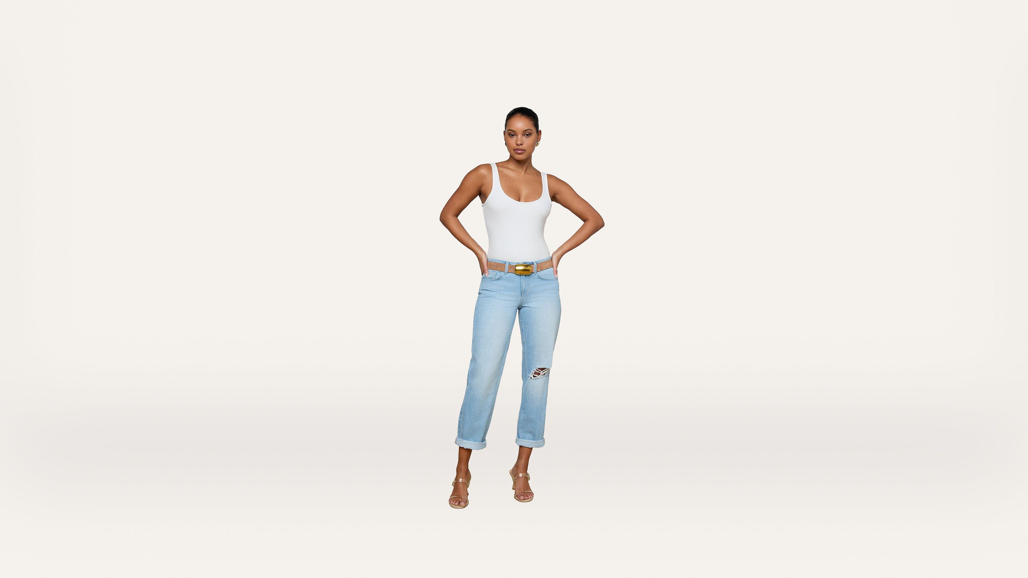 A woman stands against a plain white background, wearing a white tank top, light blue ripped jeans, a yellow belt, and heeled sandals, with her hands on her hips.