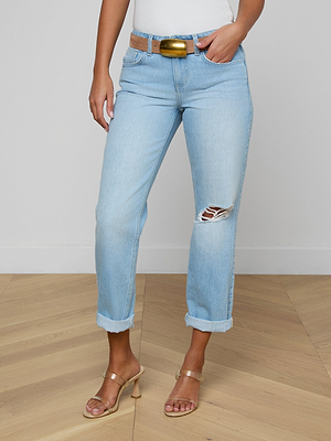 Someone in Nevia Slouchy Straight-Leg Jeans—light blue, slouch fit, rolled-up hems, rip on one knee—paired with a brown belt with a large gold buckle, white top, and clear-heeled sandals stands on a wooden floor.