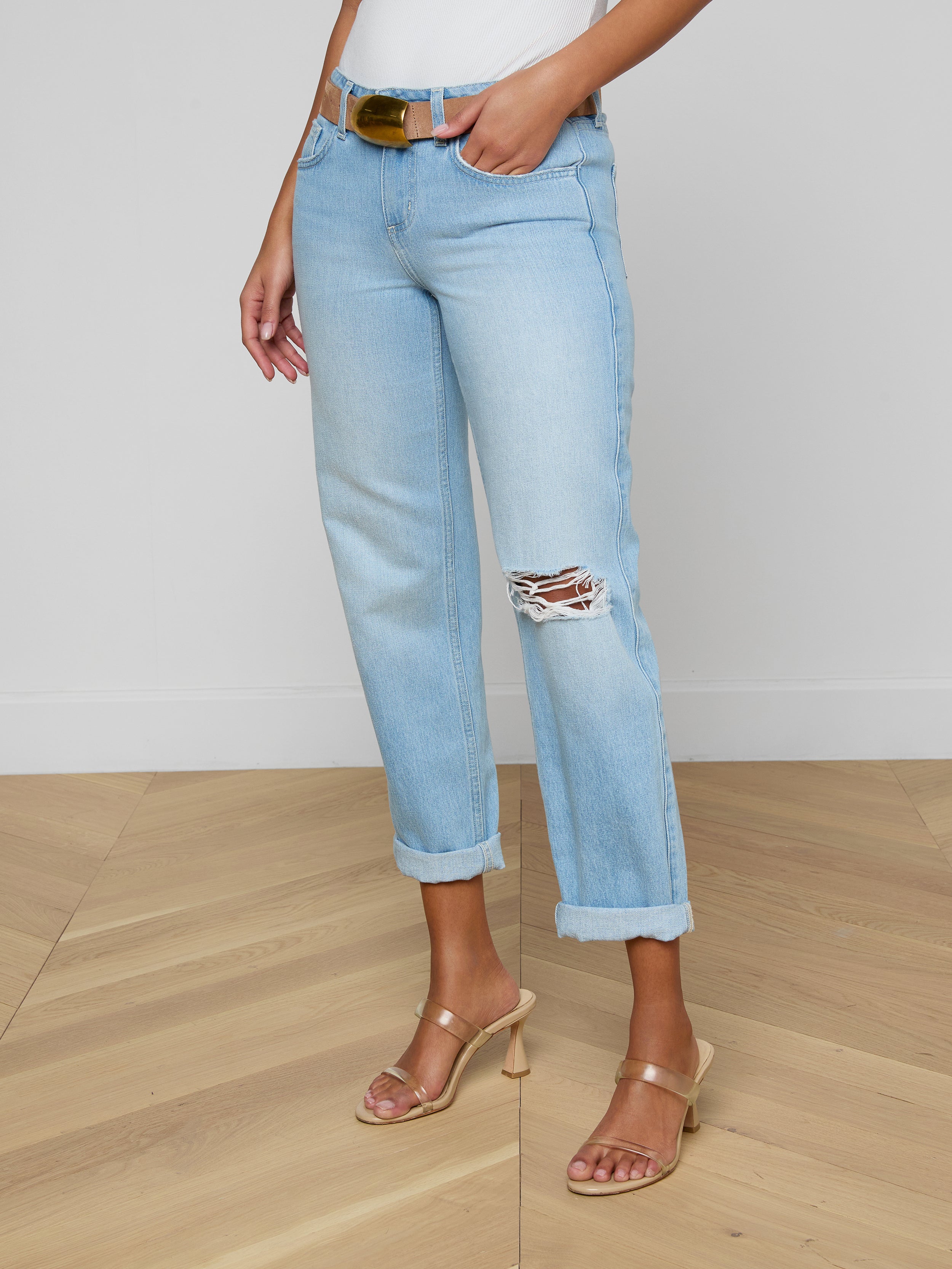 Wearing the Nevia Slouchy Straight-Leg Jean—light blue, cuffed with a ripped knee and brown belt—a person pairs them with a white top and tan heeled sandals, standing on a wooden floor against a white wall.
