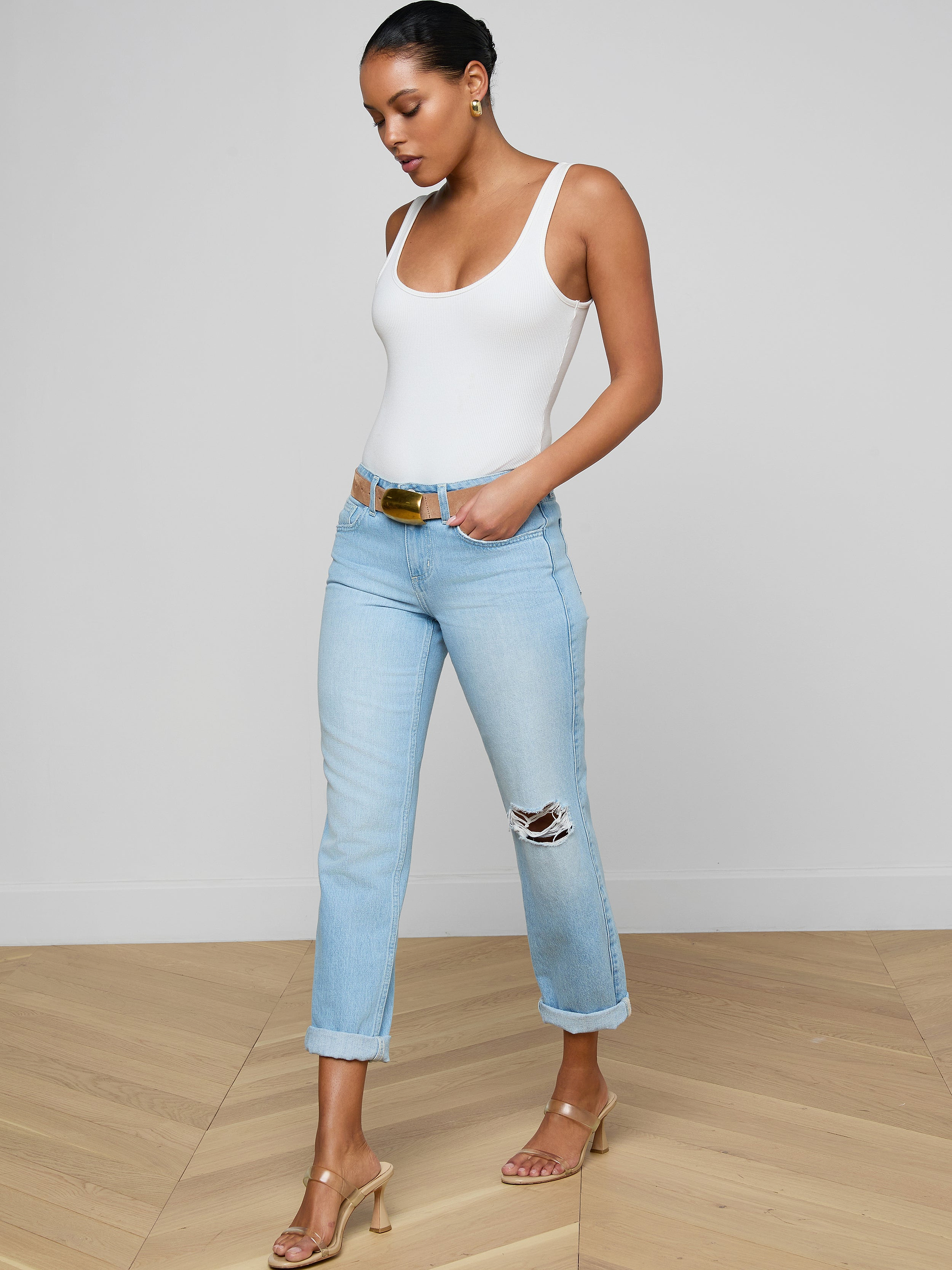 A woman stands on a wooden floor wearing a white tank top, Nevia Slouchy Straight-Leg Jean in light blue with rolled cuffs and rips, a brown belt, and nude heel sandals, looking down with one hand in her pocket.
