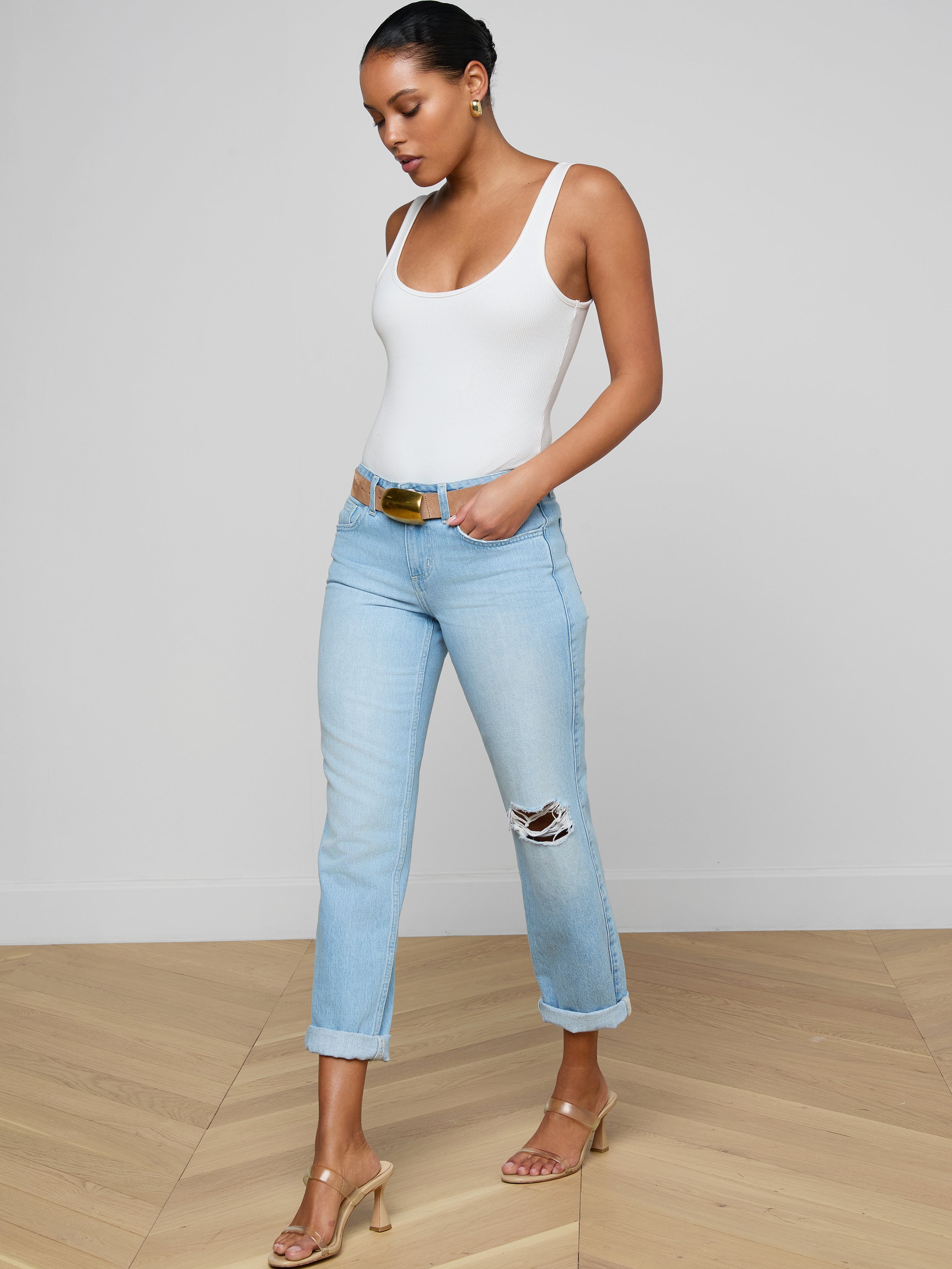 A woman stands on a wooden floor wearing a white tank top, Nevia Slouchy Straight-Leg Jean in light blue with rolled cuffs and rips, a brown belt, and nude heel sandals, looking down with one hand in her pocket.