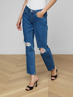 A woman wears Nevia Slouchy Straight-Leg Jeans in light wash cotton denim with black high-heeled sandals, standing on a light wood floor against a plain light background, one hand casually in her pocket.